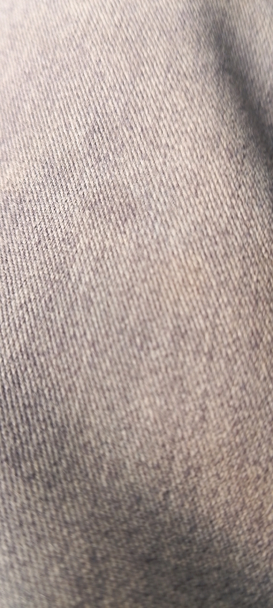 The image displays a close-up, abstract view of a textured fabric, likely denim or a similar woven material. The fabric is a muted gray-blue color, with visible diagonal lines formed by the weave of the threads. The lighting suggests a soft, diffused light source, creating subtle shadows and highlights that emphasize the texture. The focus is entirely on the fabric itself, with no discernible objects, people, or environmental context visible. There is no information in the image that could be used to determine a specific location within the city of Bekasi, Indonesia.
