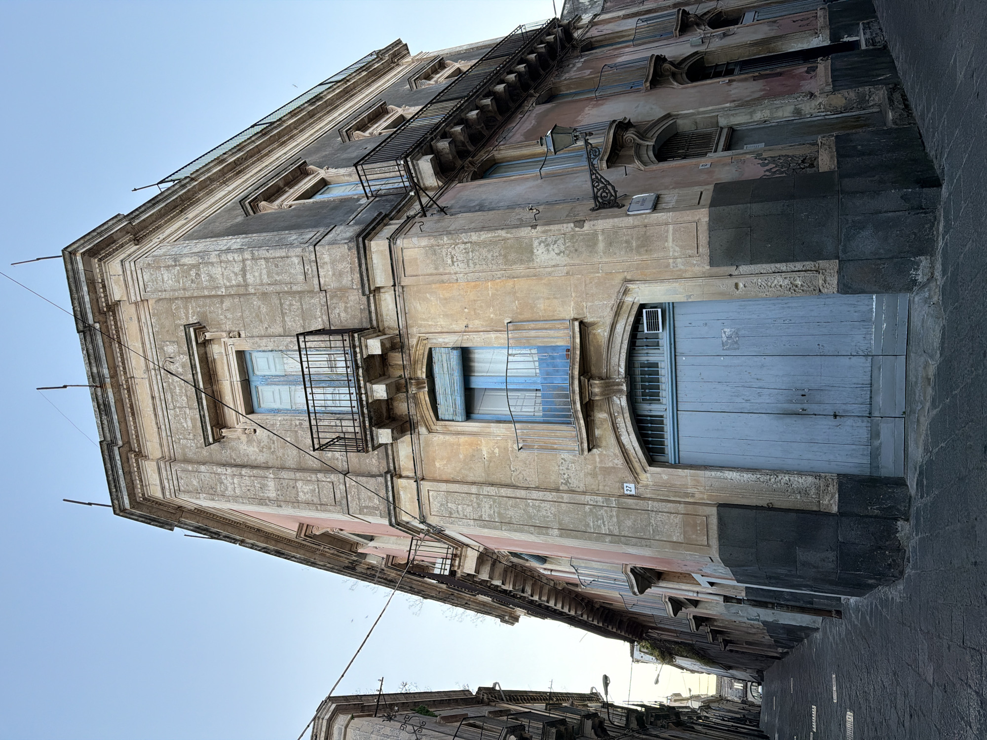 The photograph captures a street scene in Acireale, Italy, focusing on the architecture of a building along a narrow cobblestone street. The building's facade exhibits a mix of aged and weathered surfaces, with a color palette dominated by gray, beige, and hints of pastel blue on window shutters and door panels.
Architectural details include ornate window frames, small wrought-iron balconies, and a fire escape ladder clinging to the side of the building. Wires crisscross the sky above. At street level, a large blue door anchors the base of the structure. A street number '27' is visible on the facade beside the door. The perspective is from a low angle, accentuating the height of the building and its position within the tight streetscape. The sky is light blue.