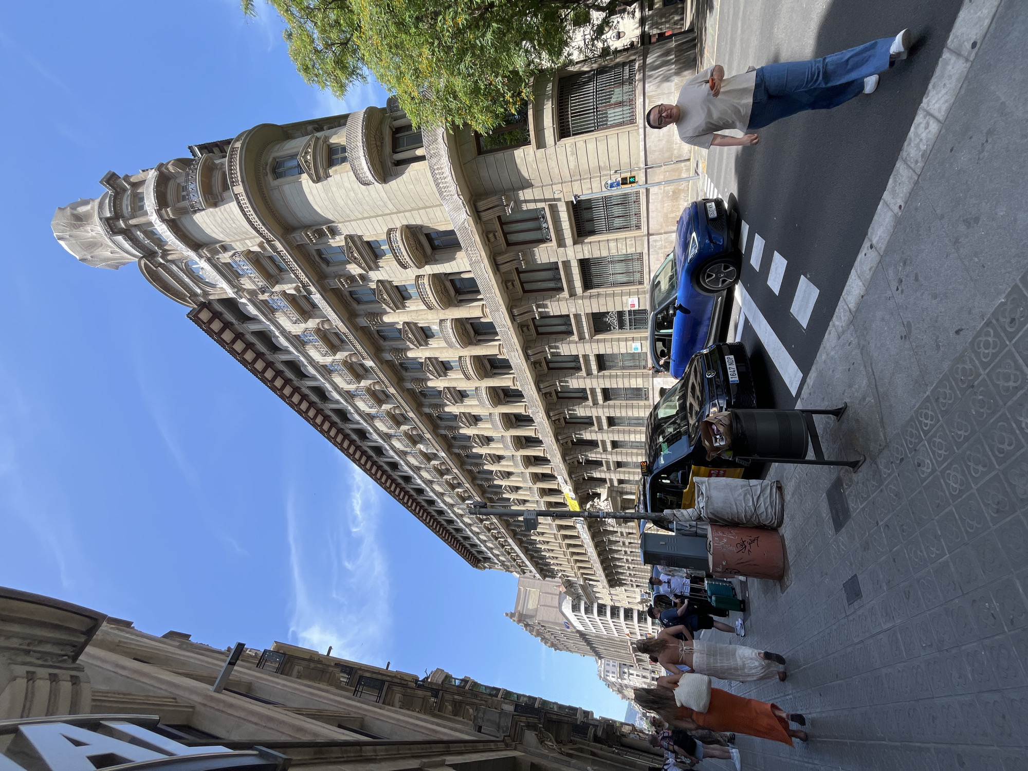 The image shows a wide street in Barcelona, Spain, with pedestrians and vehicles. The architecture features classic European buildings with balconies and ornate facades. On the right side of the street, a distinctive triangular building with a rounded corner dominates the skyline, its upper section covered by scaffolding or protective sheeting. A large green tree partially obscures the building. A taxi is parked near a crosswalk, and a blue car is also present. Several people are walking on the sidewalk and crossing the street. A woman in a light-colored shirt and blue jeans is walking towards the viewer, holding a mobile phone. Another group of women, dressed in summer clothing, are walking away from the camera. The sky is clear blue with some wispy clouds. The street and sidewalk are paved with decorative tiles. Trash receptacles and street lamps are visible along the sidewalk.