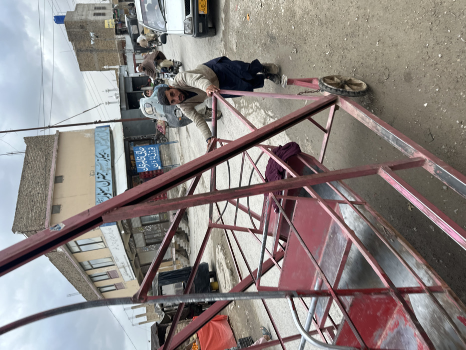 The image depicts an urban street scene in Quetta City Tehsil, Pakistan, under an overcast sky. In the foreground, a man pushes a large, disassembled, red metal structure, which appears to be a component of a carnival ride such as a Ferris wheel, equipped with several red seats. One seat contains a piece of dark red fabric. The man has a beard, wears a light-colored v-neck sweater over a dark undergarment, dark blue shalwar, and dark shoes. He is positioned behind the structure, pushing it forward with both hands.
The street surface is an uneven mixture of asphalt and dirt. To the left, a roadside stall with orange items and a red canopy is partially visible. In the mid-ground, a two-story building with a light beige facade and a brick parapet houses ground-floor businesses. A prominent sign on this building displays "بیریز جنرل سٹور" (Berez General Store) in white Urdu script on a light blue background. Below this, another blue sign reads "خانی خاطر خان فرنيش" (Khani Khatir Khan Furnish) in white Urdu script.
Vehicle and pedestrian traffic is visible in the background. A white car with a yellow license plate partially displaying "AUB-" is present on the right. Several motorcycles are on the road; one carries a man and a child, and another carries a man wrapped in a brown shawl. In the distant background, two white minarets of a mosque are discernible amidst other multi-story buildings. The overall environment suggests a bustling commercial area.