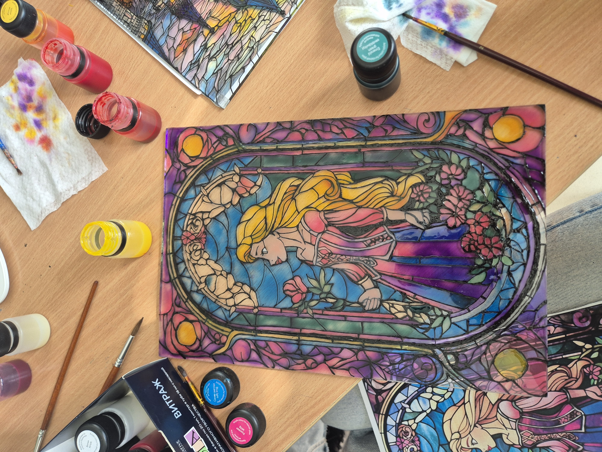 The image captures an overhead view of a wooden surface adorned with art supplies and stained-glass artworks, suggesting a creative session in an art studio or craft space.
The central focus is a rectangular piece of artwork resembling a stained-glass panel, depicting a character with flowing blonde hair, likely Rapunzel, in a pink and purple gown. She is shown in profile, holding a red rose, surrounded by various flowers and intricate, colorful patterns in blues, greens, purples, and yellows, all outlined in black to mimic traditional stained glass.
Around this main artwork, several art supplies are scattered. These include multiple small plastic bottles of vibrant liquid paints in red, yellow, green, blue, pink, and white/clear, some open and some closed. A white paper towel with colorful paint smudges lies near a small paintbrush. Other paintbrushes with wooden handles are also visible. A cardboard box for a paint set, labeled "ВИТРАЖ" (Vitrage/Stained Glass), is prominently displayed, with additional text indicating "АКРИЛОВЫЕ КРАСКИ" (Acrylic paints) and "ДЛЯ ДЕКОРАТИВНОГО ТВОРЧЕСТВА" (For decorative creativity), along with specifics like "20 мл, 1 контур в тубе 18 мл с насадкой" (20 ml pots, 1 contour tube 18 ml with nozzle). Individual paint bottles also have labels like "Белая" (White), "Розовая Rose" (Pink Rose), "Синяя светлая Blue light", and "Изумрудная Emerald green".
Partially visible at the top and bottom right are other pieces of stained-glass art, some appearing to be in different stages of completion or reference pieces. A blue denim-like fabric is seen at the bottom right. The scene depicts an artistic endeavor, likely focusing on stained-glass painting or a similar craft, with the Russian/Bulgarian text on the labels suggesting a local context, such as Pavlikeni, Bulgaria.