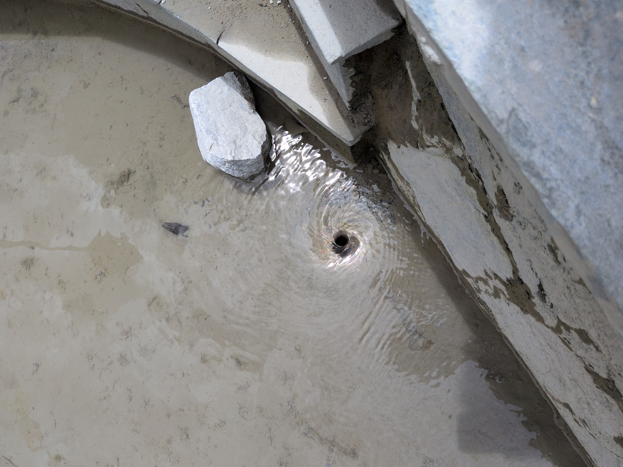 The image shows a drain where water is swirling into a funnel shape. The drain is located in a sandy area with some concrete structures around it. There is a rock to the left of the drain. The water is murky and appears to be dirty. There are no people or animals visible in the image. The location is identified as Panamik, India.