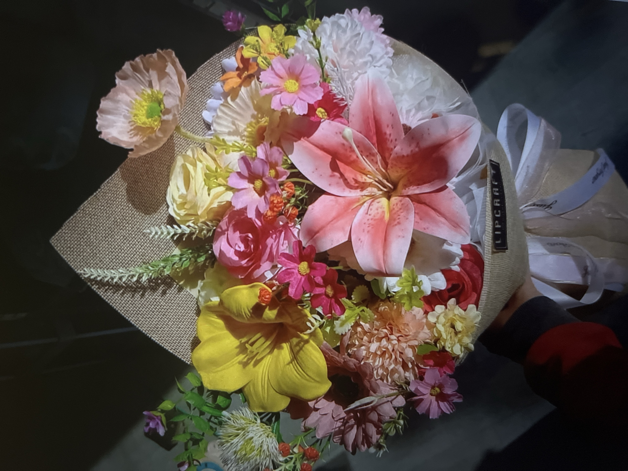 A hand holds a bouquet of artificial flowers. The bouquet is wrapped in burlap and tied with a white ribbon. It contains a large pink lily, a large yellow lily, pink roses, and various smaller pink, yellow, and orange flowers. There is also a light pink poppy flower visible at the top. The background is dark, with some light reflections. A small black tag with white lettering reading "LIPCRAFT" is attached to the burlap wrap. The scene appears to be indoors.