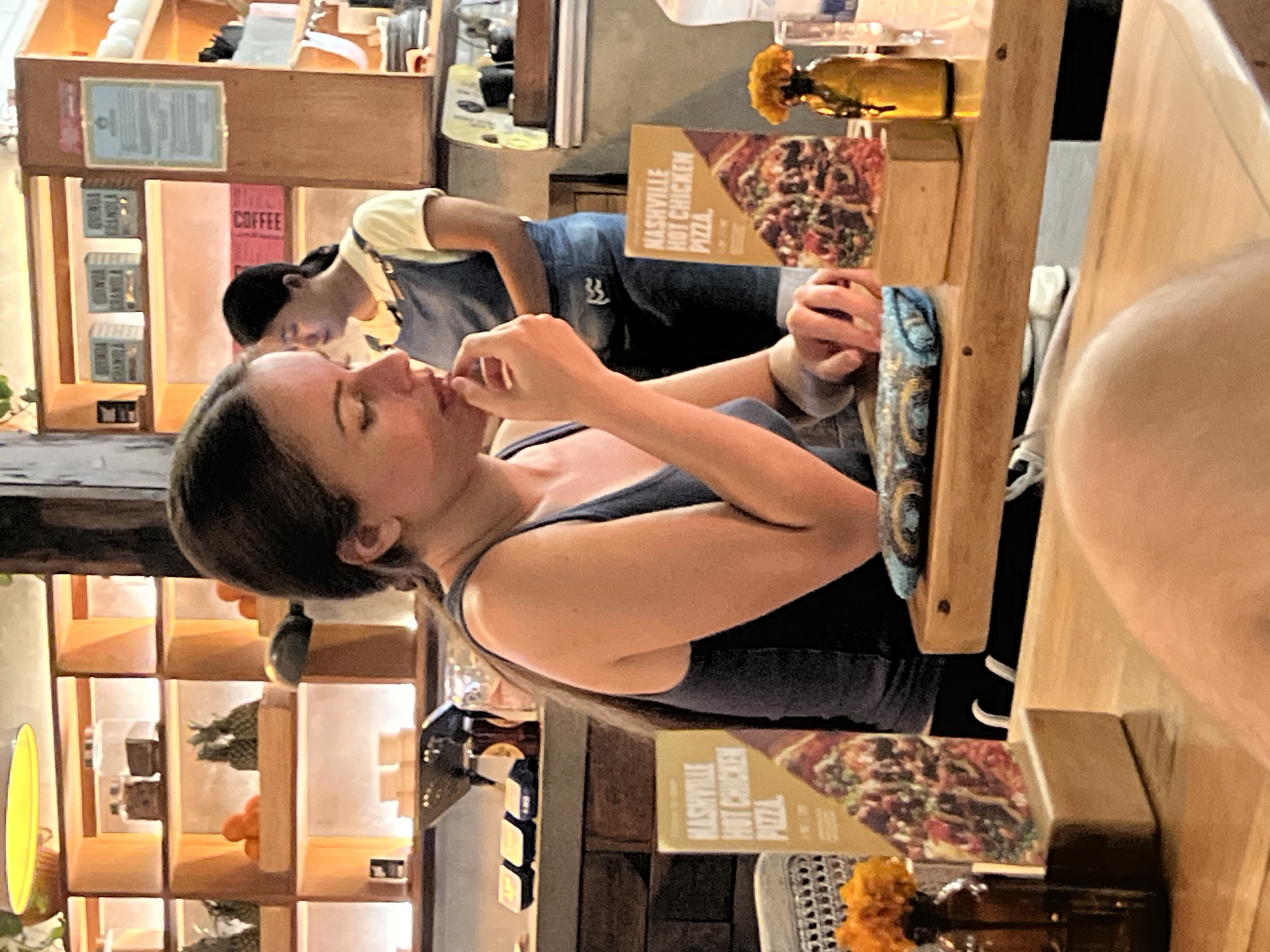 The image appears to be taken in a restaurant or cafe in North Kuta, Indonesia, as indicated by the "Nashville Hot Chicken Pizza" signage (a common menu item in such establishments). A Caucasian woman with dark hair is seen eating something with her hands. An Asian woman is partially visible in the background, wearing a yellow shirt and a denim apron, suggesting she might be a staff member. The restaurant setting includes shelving with decorative items like plants and jars. There's a rustic, wooden table with what appears to be a bottle of water and a small vase of flowers. The foreground is dominated by a person whose body is mostly out of frame.