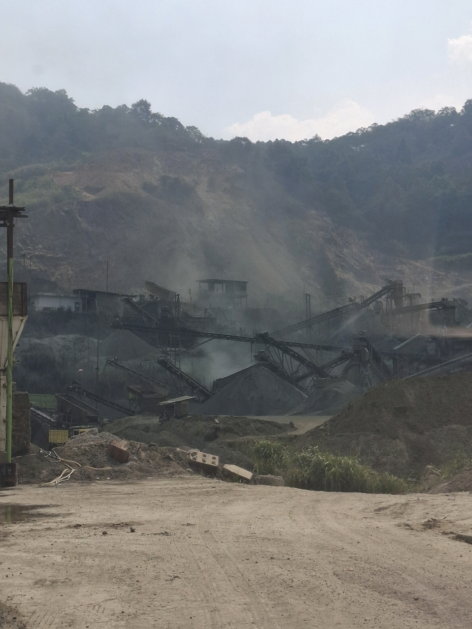 The image depicts a dusty industrial scene, likely a quarry or processing plant, situated in a hilly, vegetated environment. The foreground is a dirt track with scattered debris, including hoses and what appear to be construction materials. In the mid-ground, multiple conveyor belts and processing machinery are visible, with dust plumes indicating ongoing operations. Buildings, possibly part of the facility, are present amidst the machinery. The background features steep, eroded hillsides covered in vegetation, with a hazy sky overhead. The overall impression is one of an active industrial site in a natural, albeit disturbed, landscape. The presence of dust and industrial equipment suggests a mining or aggregate processing operation. The location is identified as Cijangkar, Indonesia, implying a potential link to the region's natural resources or industrial activities.