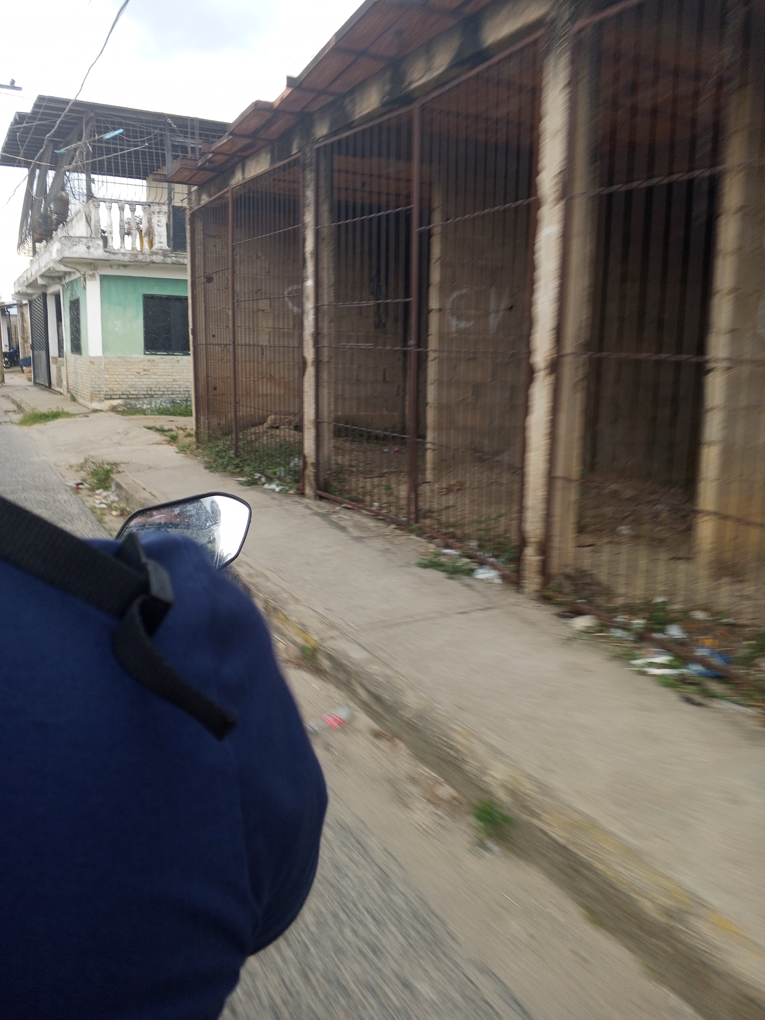 The image, taken from a moving vehicle (likely a motorcycle, given the presence of a side mirror), presents a street scene in Tinaquillo, Venezuela. The focus is on a series of barred structures running parallel to the road. These structures appear to be part of a building, possibly storage units or garages, distinguished by their aged concrete frames and heavy iron bars. Inside each barred section, the ground is visible, scattered with debris. The road itself is paved, with visible signs of wear and scattered trash along the edges. In the background, a two-story building with a light green facade and wrought iron accents is partially visible. Overhead power lines traverse the scene. The lighting suggests daytime, under overcast skies. A portion of the vehicle is visible in the foreground, showing a dark blue material and a black strap.