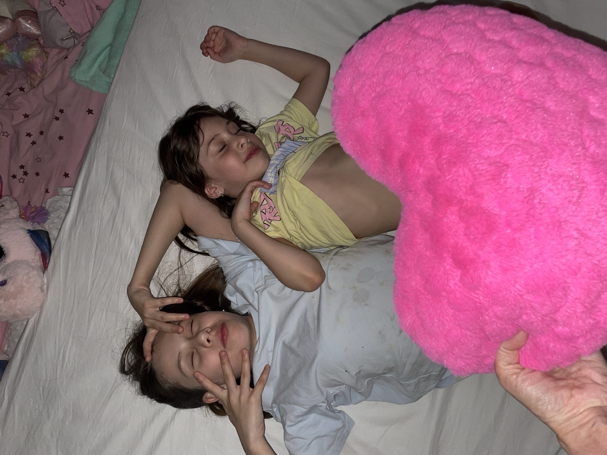 Two young girls are sleeping on a white bed with a pink heart-shaped pillow nearby. One girl, wearing a yellow t-shirt, is lying with her eyes closed, her arm raised. The other girl, wearing a light blue t-shirt, has her hand near her face. A hand holds the edge of the pink pillow in the lower right corner. Various toys and bedding are visible on the left side of the bed. The location is not discernable from the image.
