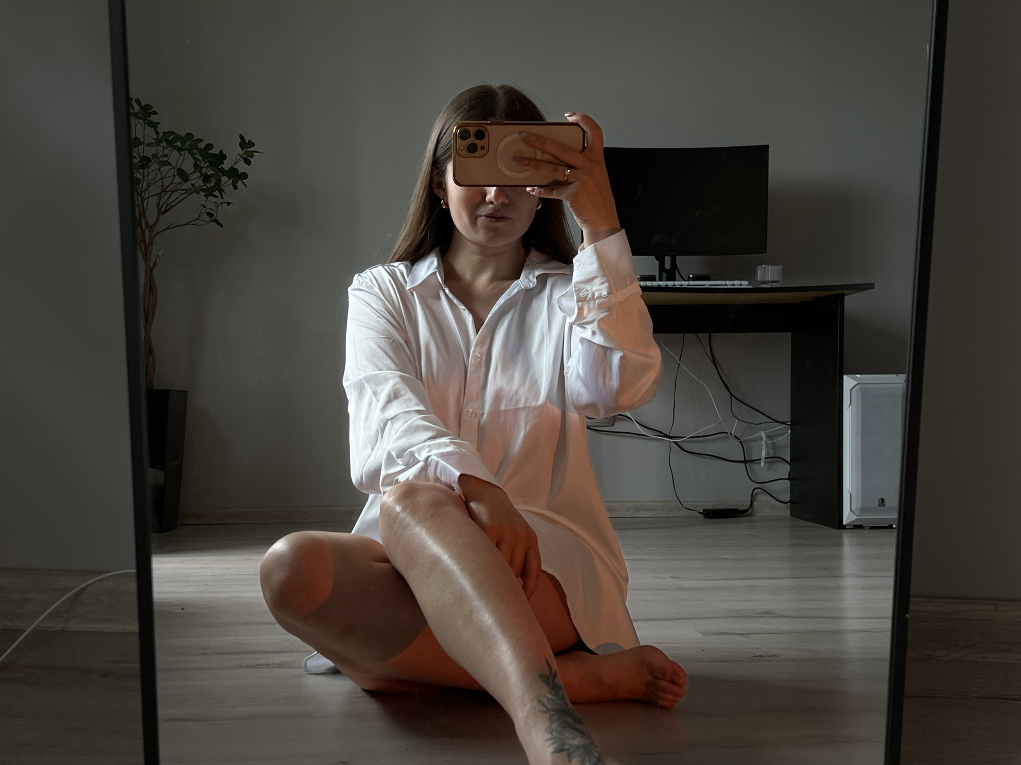 The image depicts a young adult female taking a mirror selfie indoors. She is seated on a light-colored wooden floor, facing the mirror directly. She has long brown hair parted down the middle and wears a loose-fitting, white oversized button-down shirt. Her face is largely obscured by a gold-cased smartphone, likely an iPhone with a visible MagSafe ring, which she holds with her right hand in front of her eyes. Her chin and mouth are visible, showing a subtle smile. Small gold hoop earrings are visible on her earlobes, and a ring is on her right ring finger.
Her right leg is extended forward and crossed over her left leg, which is mostly obscured. The visible right leg shows distinct tan lines on the thigh and a dark-inked tattoo on the lower shin/ankle area, appearing to be a floral or foliage design.
The background, reflected in the mirror, shows a simple, modern room. To the left, a tall potted plant with green leaves and thin branches stands against a plain light gray wall. To the right, a dark-colored desk is visible, featuring a black computer monitor, a keyboard, and a small clear object. Numerous black and white cables are visible behind and beneath the desk. A white computer tower or PC case is partially visible on the floor to the right of the desk. The walls are light gray, and a light-colored baseboard runs along the bottom. The overall lighting is subdued, with some warm light illuminating the woman's skin and shirt.
While the prompt requests the location to be Ternopil, Ukraine, the image itself does not contain any discernible visual information to confirm this specific city. The environment appears to be a residential or home office setting.