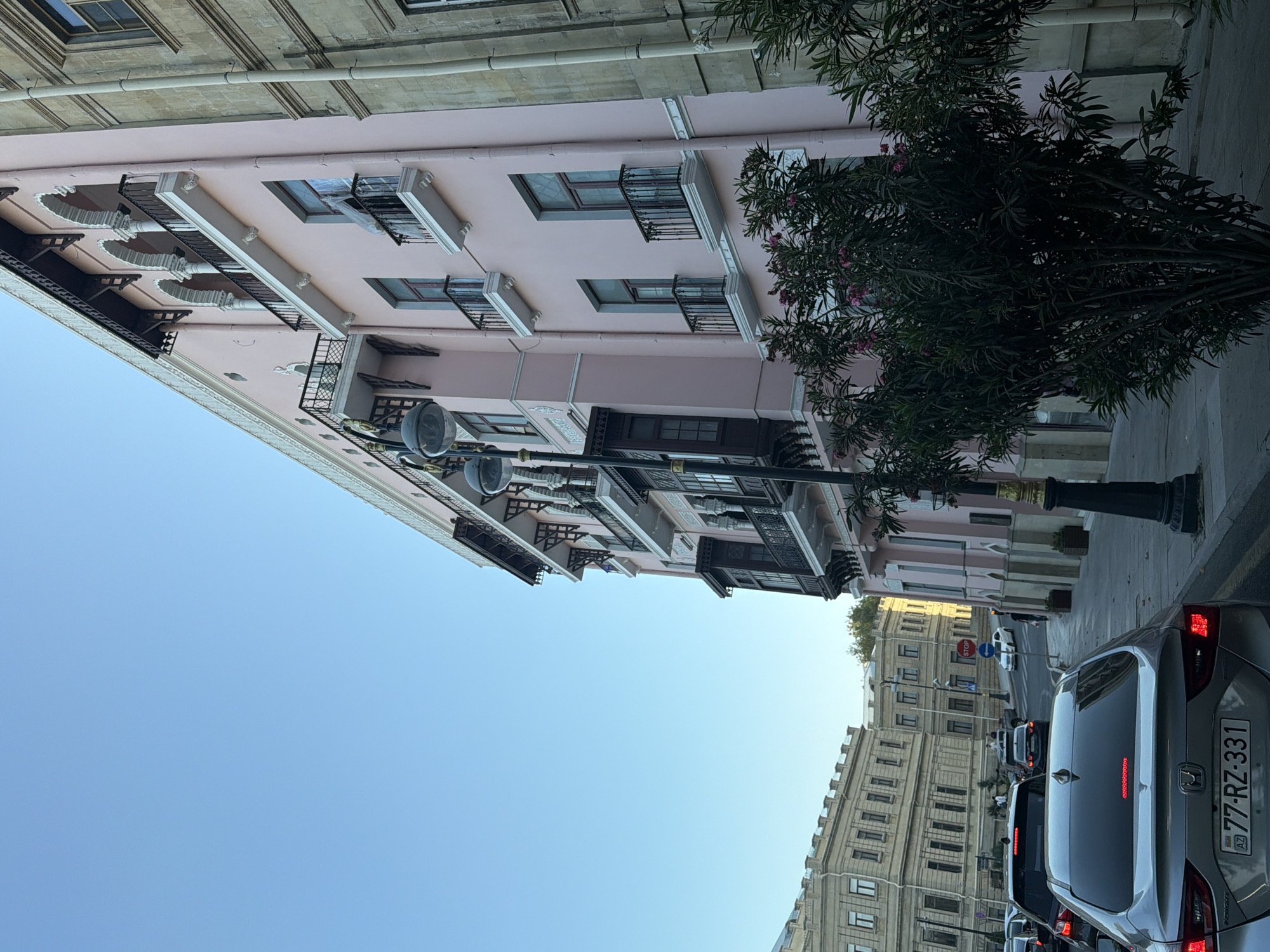 The image shows a street scene in Sabail Raion, Azerbaijan, with a light pink building dominating the right side of the frame, featuring multiple arched balconies and dark wooden window frames. To the left, a row of beige stone buildings with classical architectural details is visible. The street is occupied by several cars, indicating traffic. In the foreground, the rear of a silver car with the license plate "77-RZ-331" is prominently displayed. A street lamp with multiple globes stands near the pink building, and a dense green bush with pink flowers is in the lower right corner. The sky is clear and blue, suggesting daylight.