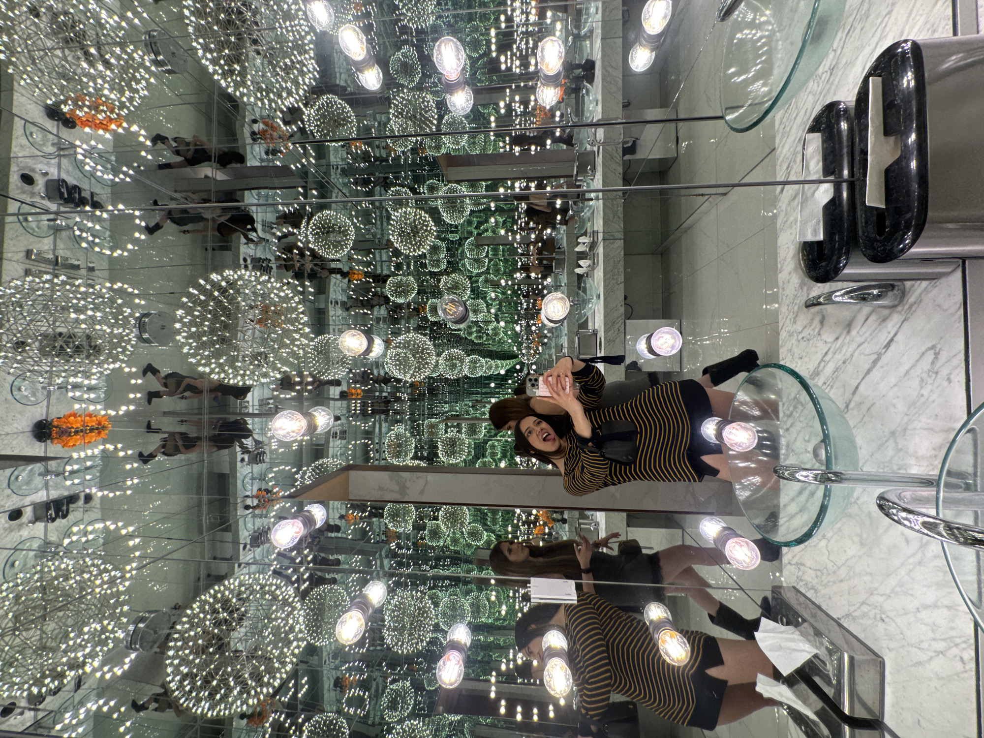 A young woman in a striped dress and hat takes a selfie in a mirrored room with multiple reflections of herself and the environment. The room appears to be a bathroom with glass bowl sinks, paper towel dispensers, and decorative lighting fixtures, including orbs resembling dandelions. The mirrors create an "infinity room" effect. The image was taken in Puebla, Mexico.