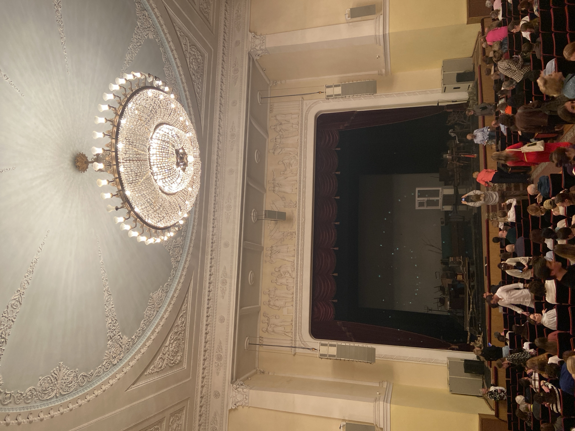 The image shows an audience in an ornate theatre. The room has a large, decorative chandelier hanging from a light blue ceiling. The walls are cream-colored and decorated with classical figures. A stage with a red curtain is visible in the background. The audience is seated in red chairs. The location is likely a theatre in the city of Sergiyev Posad, Russia.
