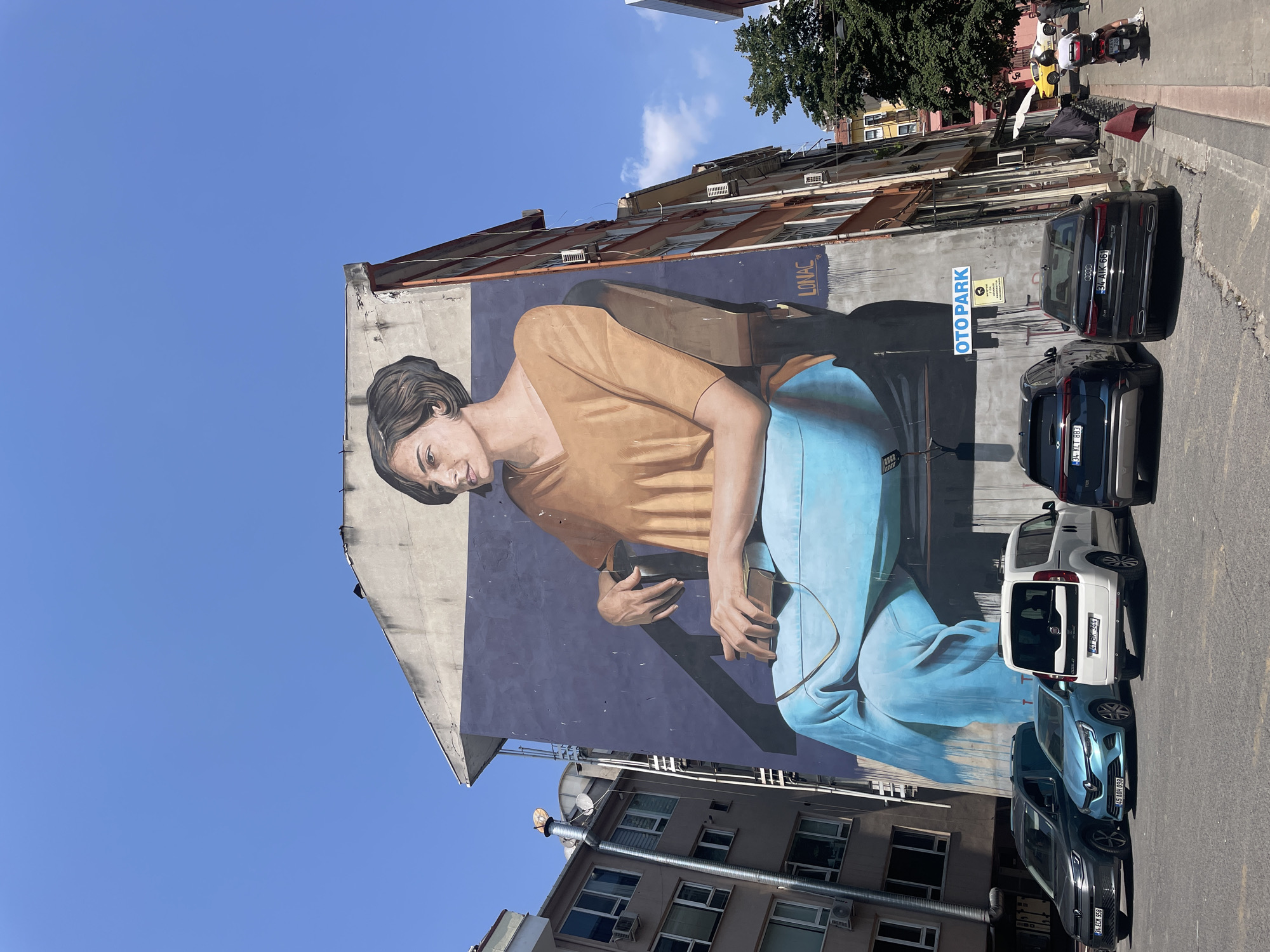 The image shows a large mural painted on the side of a building in Kadıköy, Turkey. The mural depicts a woman sitting in a chair, wearing a tan shirt and blue pants, with her legs crossed. She is holding a book or phone in her hands. The mural is vibrant and colorful, with the woman's face and clothing rendered in realistic detail. The building itself is multi-story and appears to be an apartment or office building. In front of the building, there are several parked cars, including a white van, a blue SUV, and a black sedan. A street sign that reads "OTO PARK" is visible on the side of the building, indicating a parking area. The street is paved and there are sidewalks on either side. The sky is clear and blue. The overall scene is one of an urban environment with a prominent piece of street art.