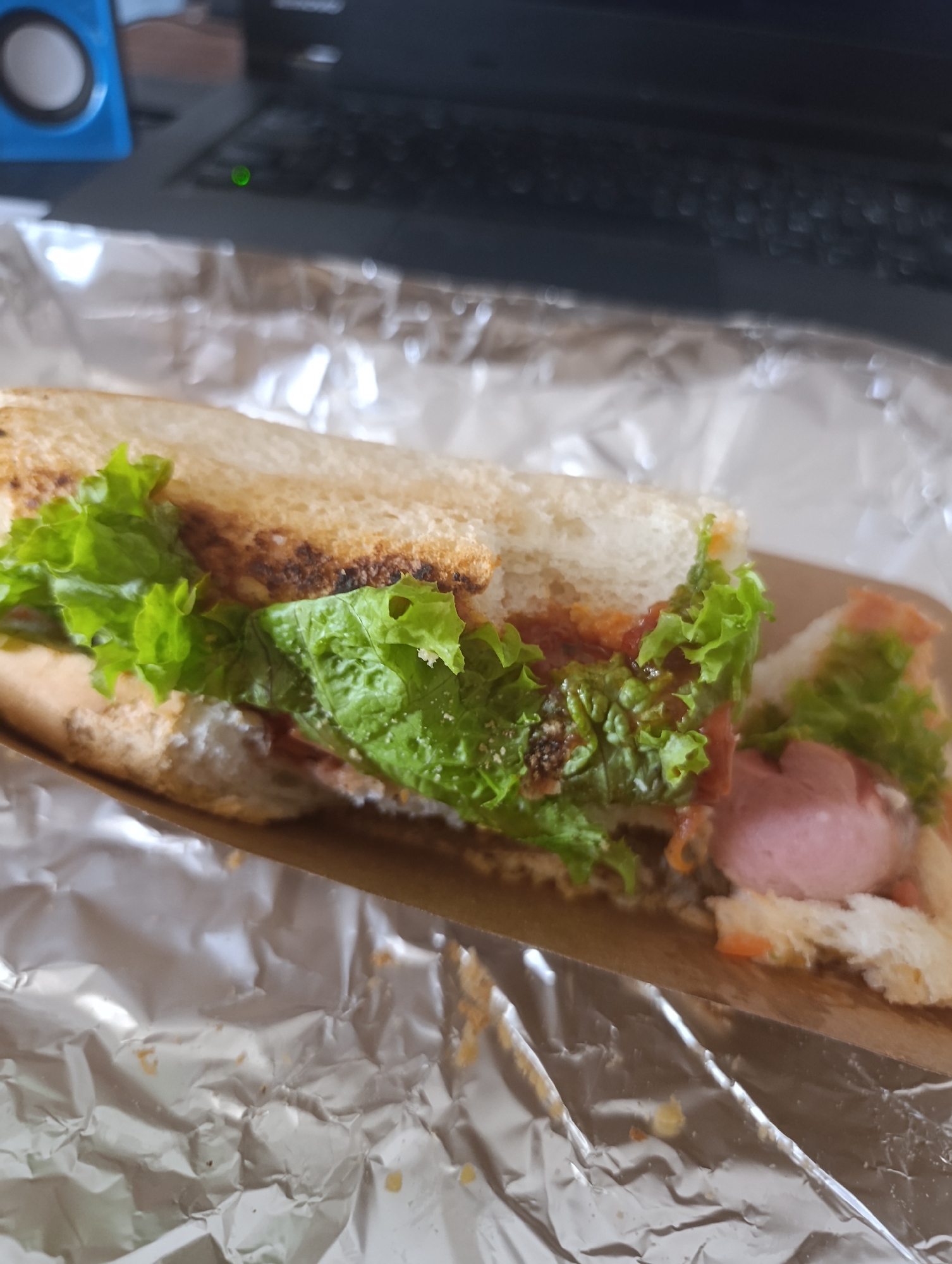 A close-up shot shows a sandwich on foil, with a blurred background of a laptop and a blue speaker. The sandwich appears to be made with white bread, lettuce, and a pink sausage. The foil is crumpled and reflects light. The scene is likely indoors, possibly on a desk or table, given the presence of the laptop. The location within the city of Vyshneve, Ukraine, is not discernible from the image.
