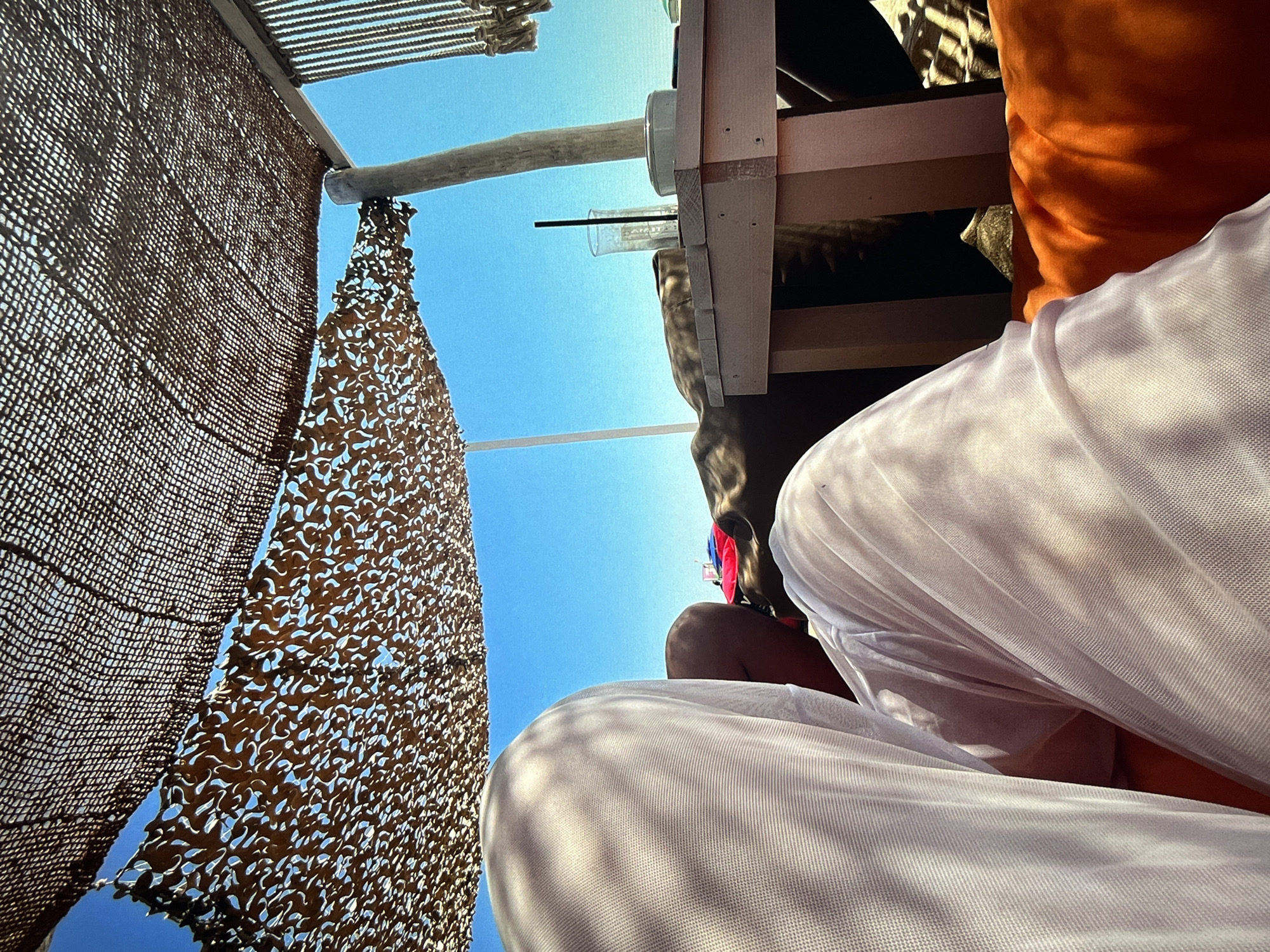 The image shows a low-angle perspective, looking upwards towards a partly obstructed sky of vivid blue. In the foreground, a large area is taken up by what appears to be white, lightly textured fabric, possibly clothing or a sheet. Moving upwards, an orange cushion is visible to the right. A pale, wooden table structure with a clear glass containing a straw is also within view. Overhead, there is a canopy or awning constructed of wooden beams and a coarsely woven material that filters the sunlight. To the left, a section of netting or camouflage netting hangs against the clear blue sky.