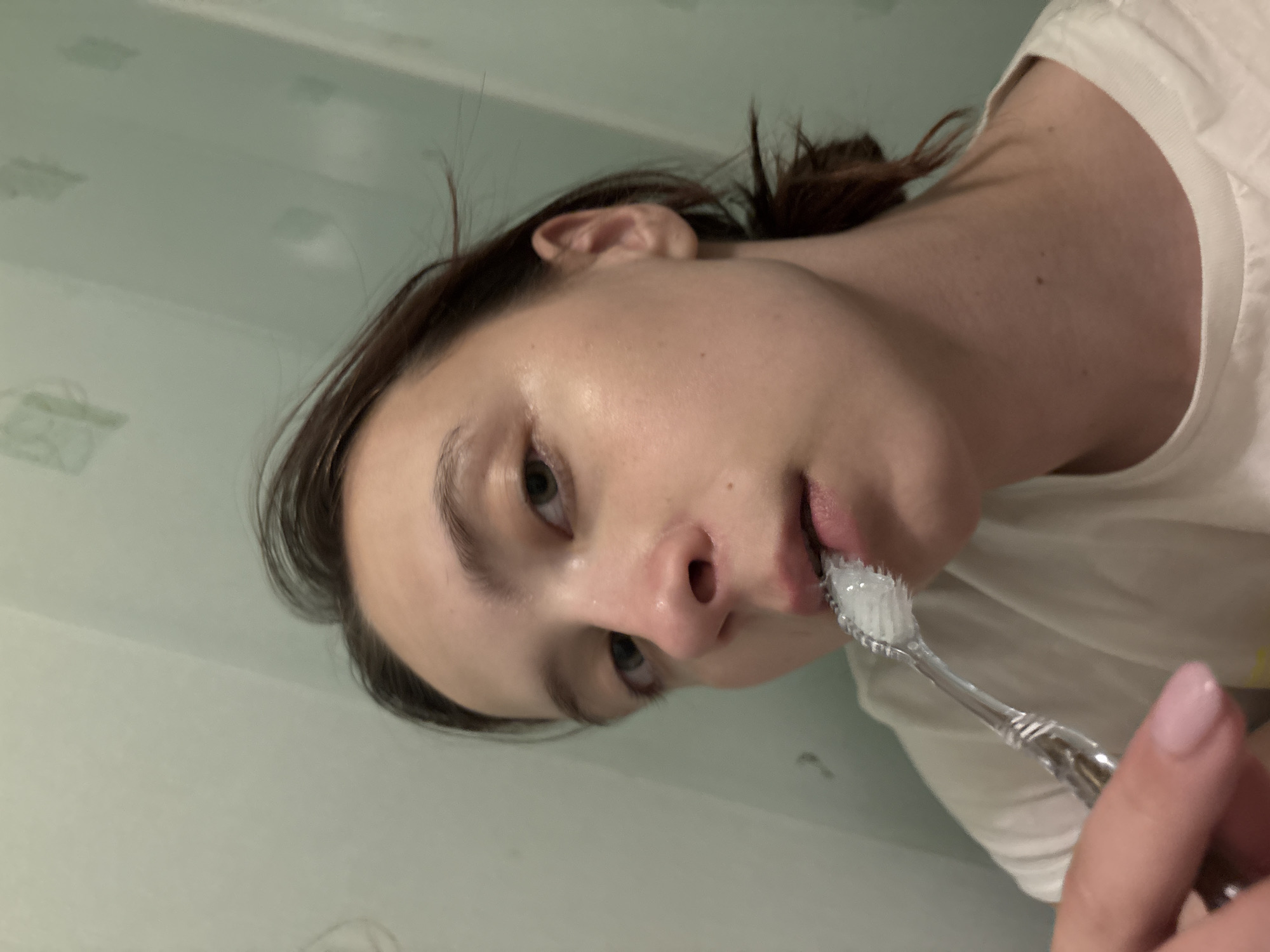 A woman is brushing her teeth. She has fair skin, dark hair pulled back, and is wearing a white t-shirt. She is holding a toothbrush with white toothpaste on it. The background is a light green wall with patterned tiles. The location is the city of Прикамский, Russia.