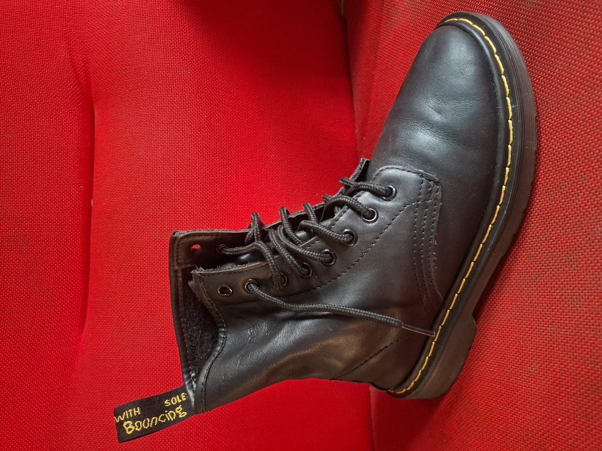A black Dr. Martens boot is photographed against a textured red background. The boot is upright, with its laces loosely tied. A yellow and black tag is attached to the top rear of the boot, reading "With Bouncing Soles". The boot exhibits typical Dr. Martens features, including yellow stitching around the sole. The background is a solid red fabric, appearing to be a chair or sofa. The image is well-lit, with the boot casting a subtle shadow. There is no information available regarding the specific location in Babakan Madang, Indonesia.