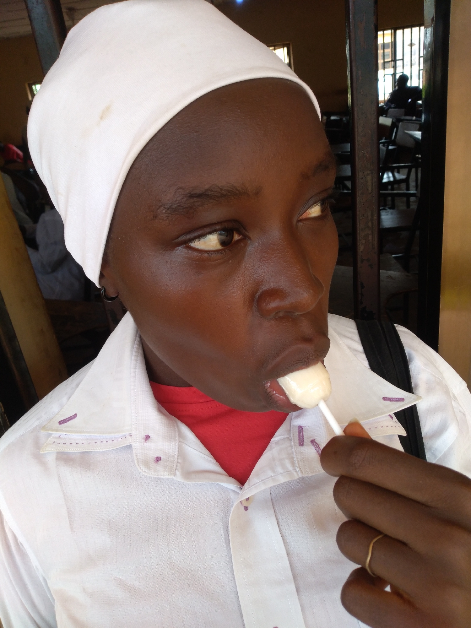 A young person, wearing a white headscarf and a white collared shirt over a red t-shirt, is holding a white lollipop to their mouth and licking it. They are looking off to the right. The scene appears to be indoors, possibly a classroom or community hall, with wooden chairs and tables visible in the background through a metal grate. The environment suggests a location within the city of Jos, Nigeria.