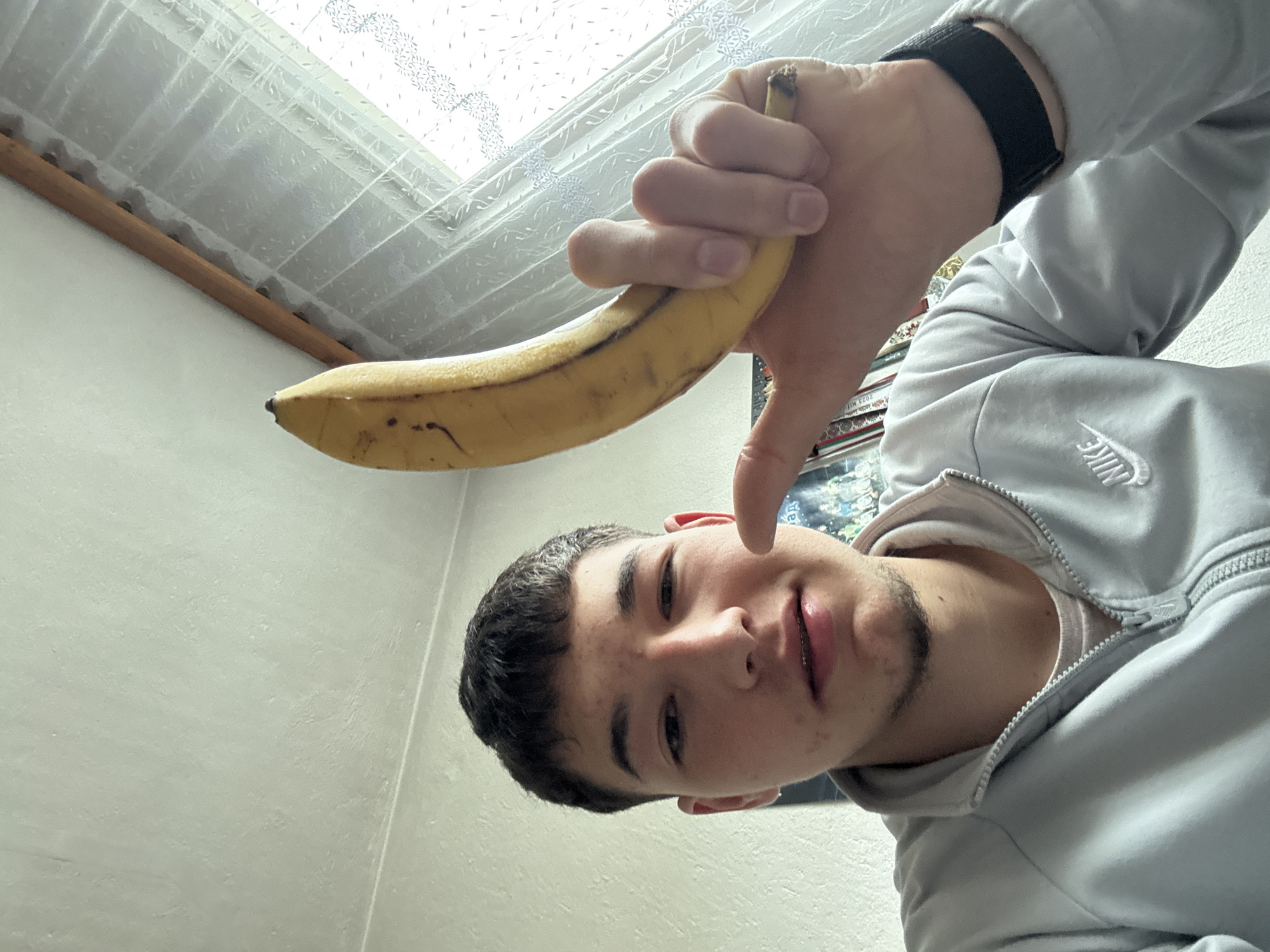 A young man is holding a banana in his hand. The photo is taken from a low angle, looking up at the man. The background includes a window with a sheer curtain and a wall. The man is wearing a grey Nike zip-up jacket and a black wristwatch. The environment is indoors. It is not possible to confirm if the photo was taken in Zlatograd, Bulgaria.