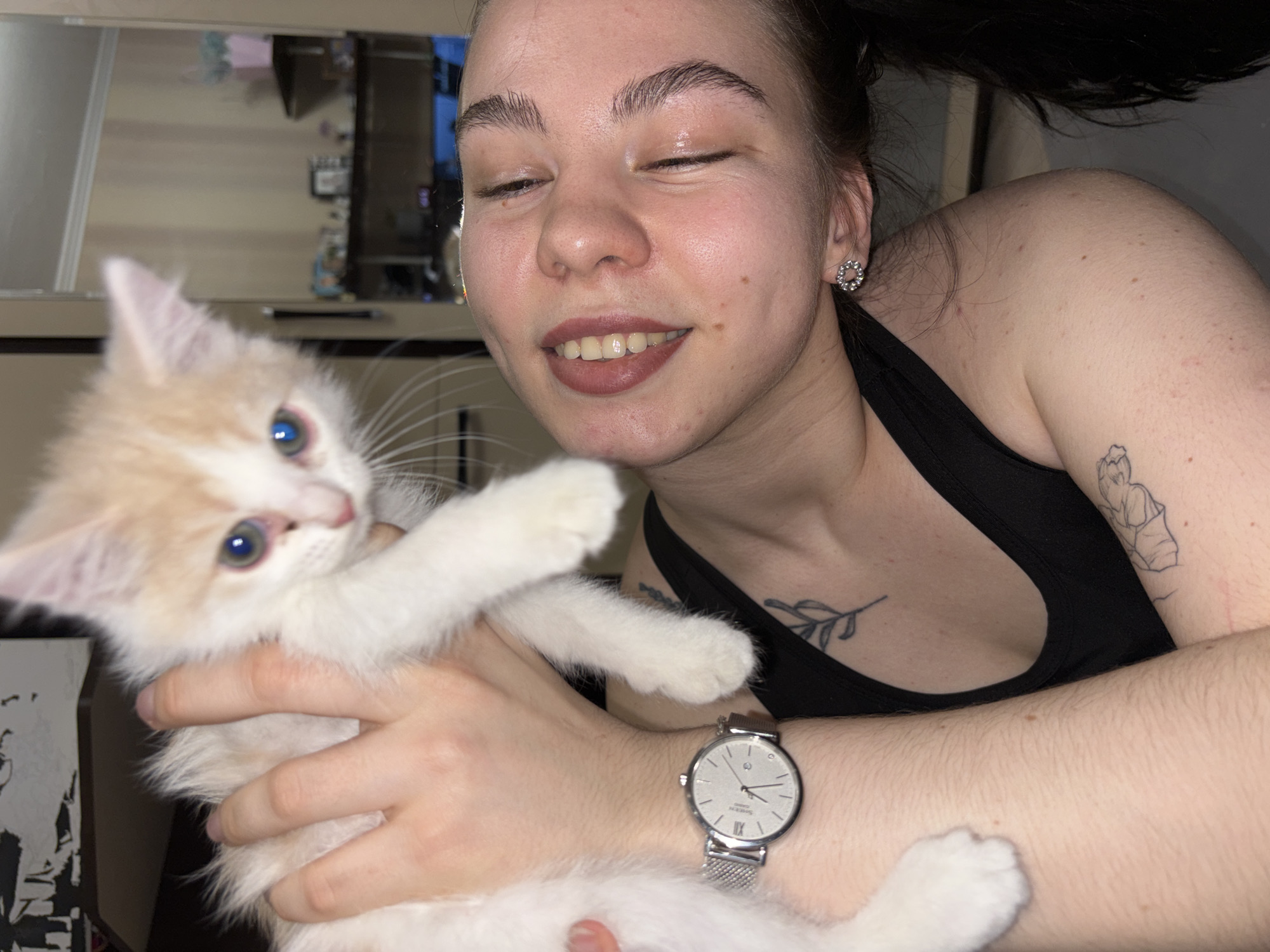 An indoor, close-up photograph captures a young woman joyfully embracing a small kitten, likely in Perm, Russia. The woman, positioned on the right side of the frame, smiles broadly with her eyes closed, showing clear contentment. She has dark hair, partially visible and pulled back, and wears a simple black tank top. Notable details on her include a small, minimalist tattoo of a leafy branch on her upper chest and a larger, uncolored outline tattoo on her upper left arm, possibly depicting a figure. She wears a circular, studded silver-colored earring in her right ear and a silver-toned watch with a mesh band on her left wrist. The watch face clearly displays "SUPER-DUPE," "QUARTZ," and Roman numerals for 3, 6, 9, and 12.
Cradled in the woman's arms is a fluffy kitten, predominantly white with light orange or cream patches, particularly on its head and back. The kitten has striking bright blue or green eyes and appears engaged, looking directly towards the viewer with its front paws raised.
The background is dimly lit and features reflections, likely from a mirror or a window, showing indistinct interior elements such as furniture or shelving units. These reflections hint at an indoor setting, possibly during the evening or night. The overall mood of the image is warm and affectionate, highlighting the bond between the woman and her pet.