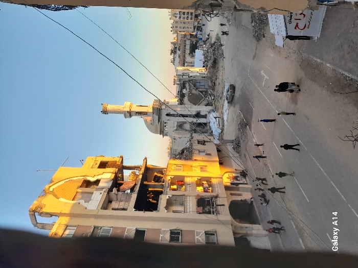 The image captures an aerial view of a war-torn city. Buildings are heavily damaged or destroyed, revealing their interiors. A mosque stands amidst the ruins. People walk along a street marked with lane dividers. Rubble and debris are scattered around the scene. A banner hangs on a wall. The sky is blue, suggesting it's daytime. "Galaxy A14" is printed on the bottom right.