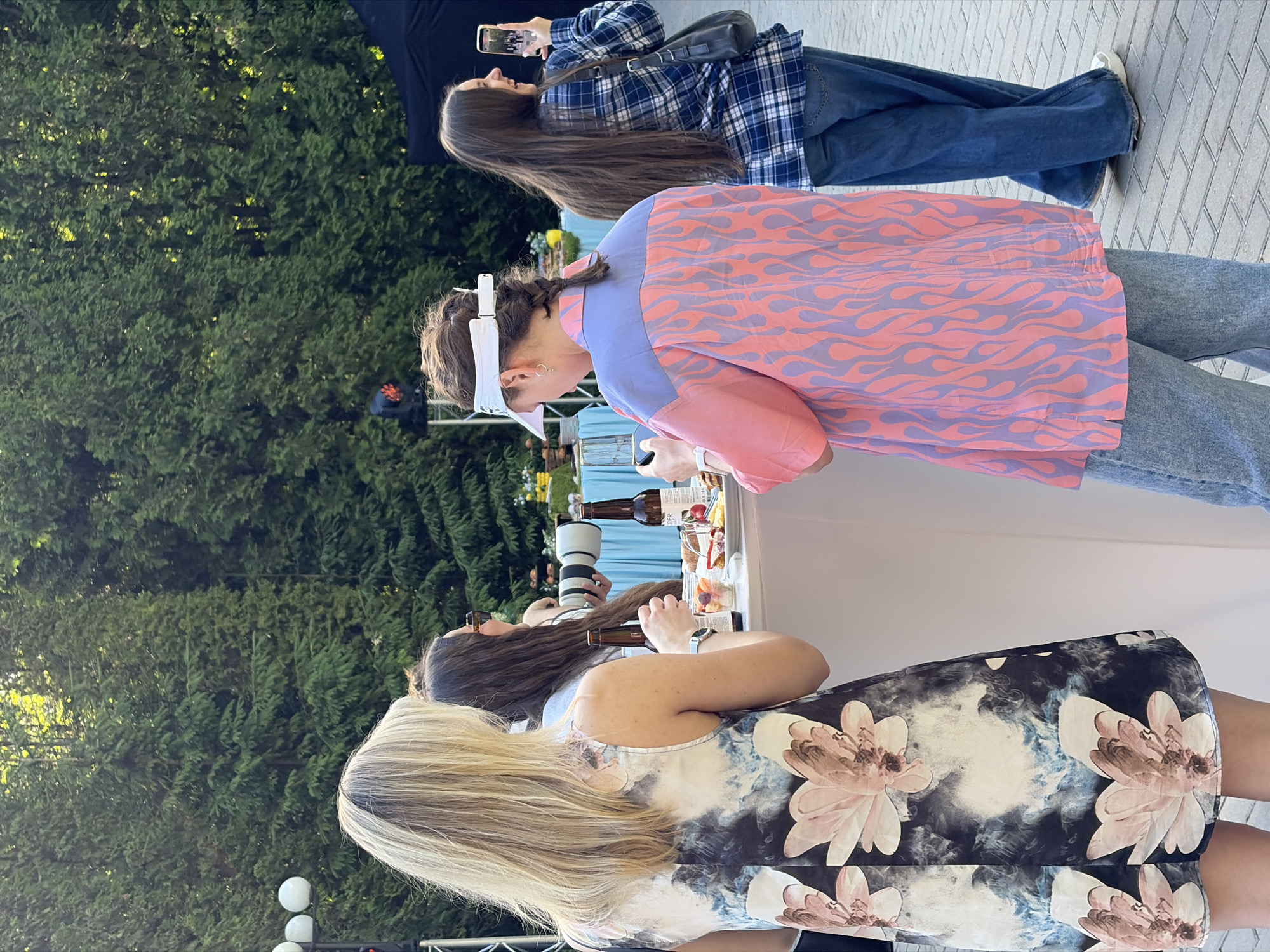 In the city of Zhdanovichi, Belarus, an outdoor social gathering is taking place with several people interacting around a standing table. A woman with blonde hair, seen from the back, wears a dark dress with a floral pattern. Next to her, another woman with long brown hair is holding a professional camera with a large lens. A third individual, facing away from the camera, sports a distinctive pink and lavender shirt with a flame pattern, paired with light-wash jeans. This person is holding a smartphone. Across from them, a fourth woman with long dark hair, wearing a blue plaid shirt and jeans, is also holding a smartphone and appears to be taking a photo or video. The table is laden with food items, including fruit and pastries, along with bottles of beverage. The background consists of lush green trees, suggesting a park or garden setting. The ground is paved with light-colored bricks.