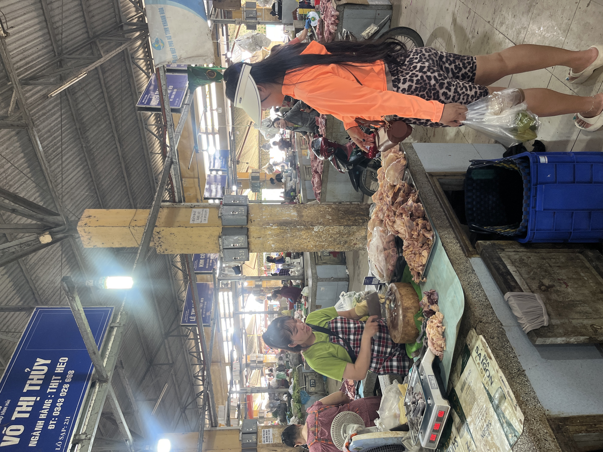 The image depicts a bustling indoor market scene in Bac Nha Trang Ward, Vietnam. In the foreground, a woman wearing a green t-shirt and a plaid apron is actively chopping meat on a large wooden block with a cleaver. To her right, another woman, dressed in an orange long-sleeved top and leopard-print shorts, stands holding a plastic bag containing drinks and what appear to be green fruits. She is looking away from the meat stall.
The market stall itself is laden with raw meat products, including poultry, displayed on trays and in plastic bags. A digital scale is also visible on the counter. In the background, other market-goers and vendors can be seen amidst more stalls and raw meat displays. The market is covered by a corrugated metal roof supported by yellow pillars, with overhead lighting illuminating the space. Several signs with Vietnamese text are visible, including one prominently displaying "VÕ THỊ THỦY" and information related to "NGÀNH HÀNG : THỊT HEO." The overall environment suggests a typical Vietnamese wet market.