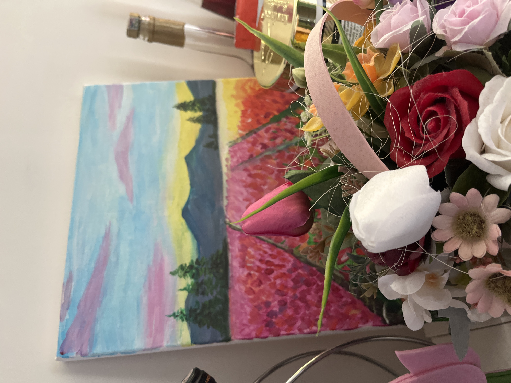 A still-life composition. In the foreground, a bouquet of artificial flowers with roses in various colors (red, white, pink), and small daisies. A light pink ribbon is interwoven in the arrangement. A painting, depicting a landscape with mountains, trees, and a field of flowers, rests on the left. The color palette of the painting is pastel. A wine bottle is partially visible above the painting. The location appears to be indoors, in Pokrovnik, Bulgaria.