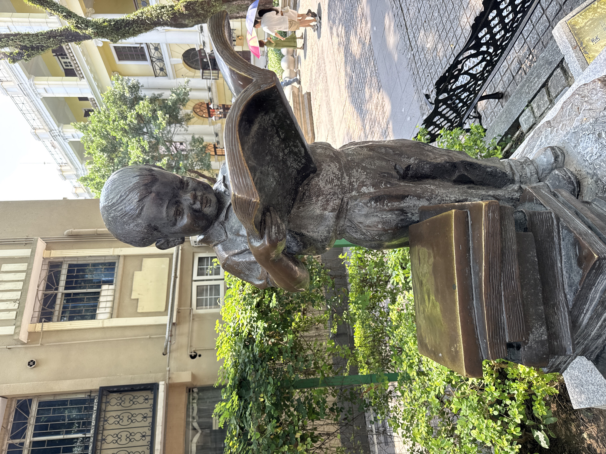 The image depicts a bronze statue of a child reading a book, situated outdoors. The statue is positioned atop a stack of books. A large book is open in front of the child, obscuring a portion of their face. In the background, buildings are visible, one a light yellow building with columns. A wrought-iron bench sits to the right of the statue. Lush green vegetation grows around the base of the statue and in the surrounding area. People are faintly visible in the background on the sidewalk. Based on the metadata and statue description, it is located in the Liwan District of China.
