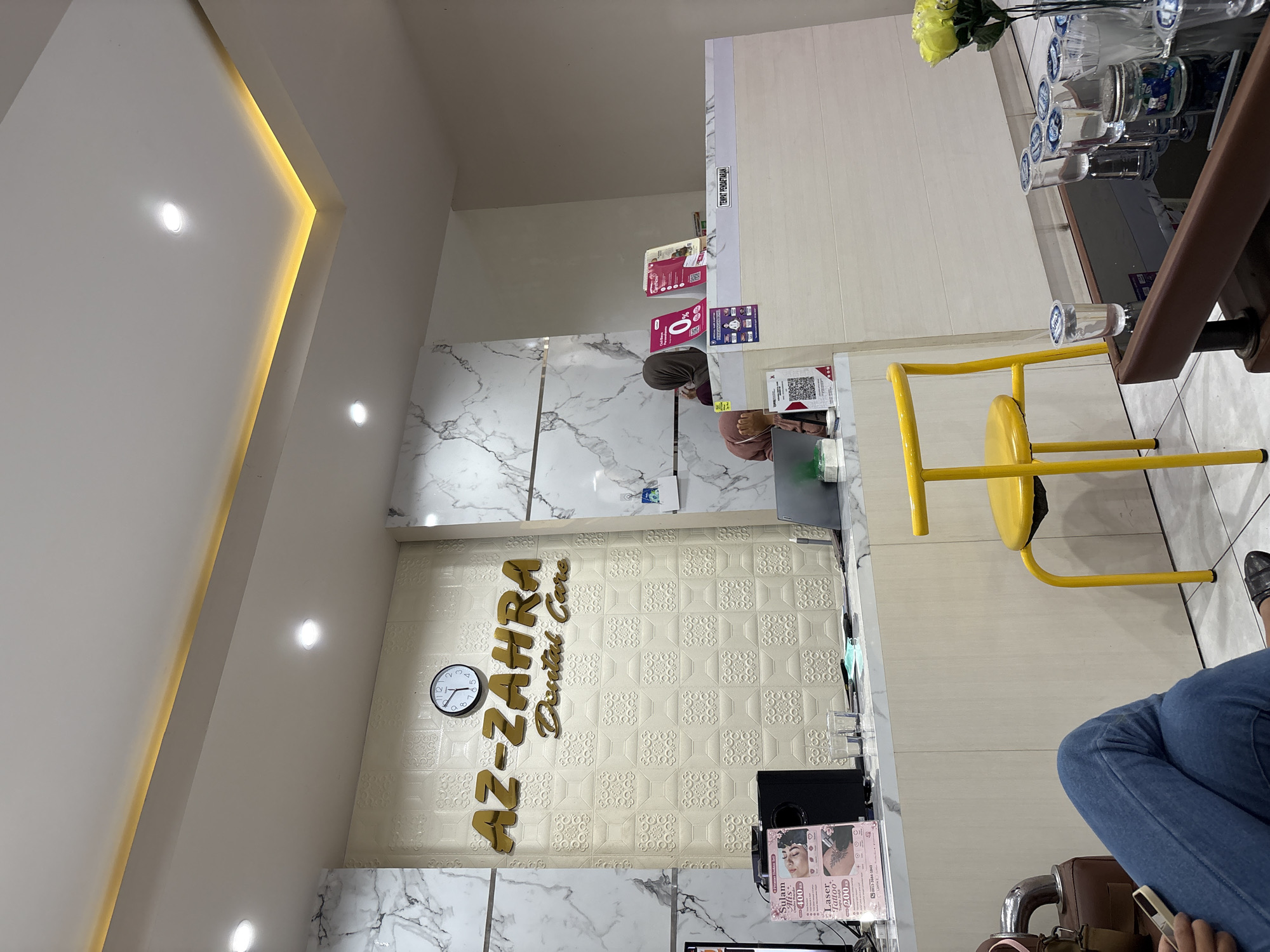 This image captures the reception or waiting area of a dental clinic named "AZ-ZAHRA Dental Care" in Kaliwates, Indonesia. The indoor scene features a modern and clean aesthetic with white walls, recessed ceiling lights, and warm strip lighting along a ceiling recess.
In the foreground, a person's lower body is visible, dressed in blue jeans, seated in a waiting area with a brown bag or purse next to them. Their hand is holding a phone, suggesting they are occupied while waiting. A bright yellow chair, possibly for another patient, is positioned near a low table. On this table, there's a vase with yellow flowers and several small plastic cups, some filled with water, arranged neatly.
The reception counter dominates the midground. It features a sleek white surface with marbled patterns on the sides, contrasting with the main wall. Behind the counter, a person wearing a head covering is partially visible, likely a receptionist. The counter partition has a sign that reads "TEMPAT PENDAFTARAN" (Registration Place). Various promotional materials and QR codes are displayed on the counter and walls.
A prominent feature is the clinic's golden-lettered name, "AZ-ZAHRA Dental Care," against a textured, light-colored wall with a repeating geometric pattern. A circular wall clock displaying approximately 12:30 is mounted to the left of the clinic's name. On the counter in the foreground, a flyer or brochure is visible, advertising "Sulam Alis 400" (Eyebrow Embroidery) and "Laser Tattoo 200," along with a phone number (0813-3604-1843). The figures likely refer to prices in hundreds of thousands (e.g., IDR 400,000). The overall ambiance is bright and professional.