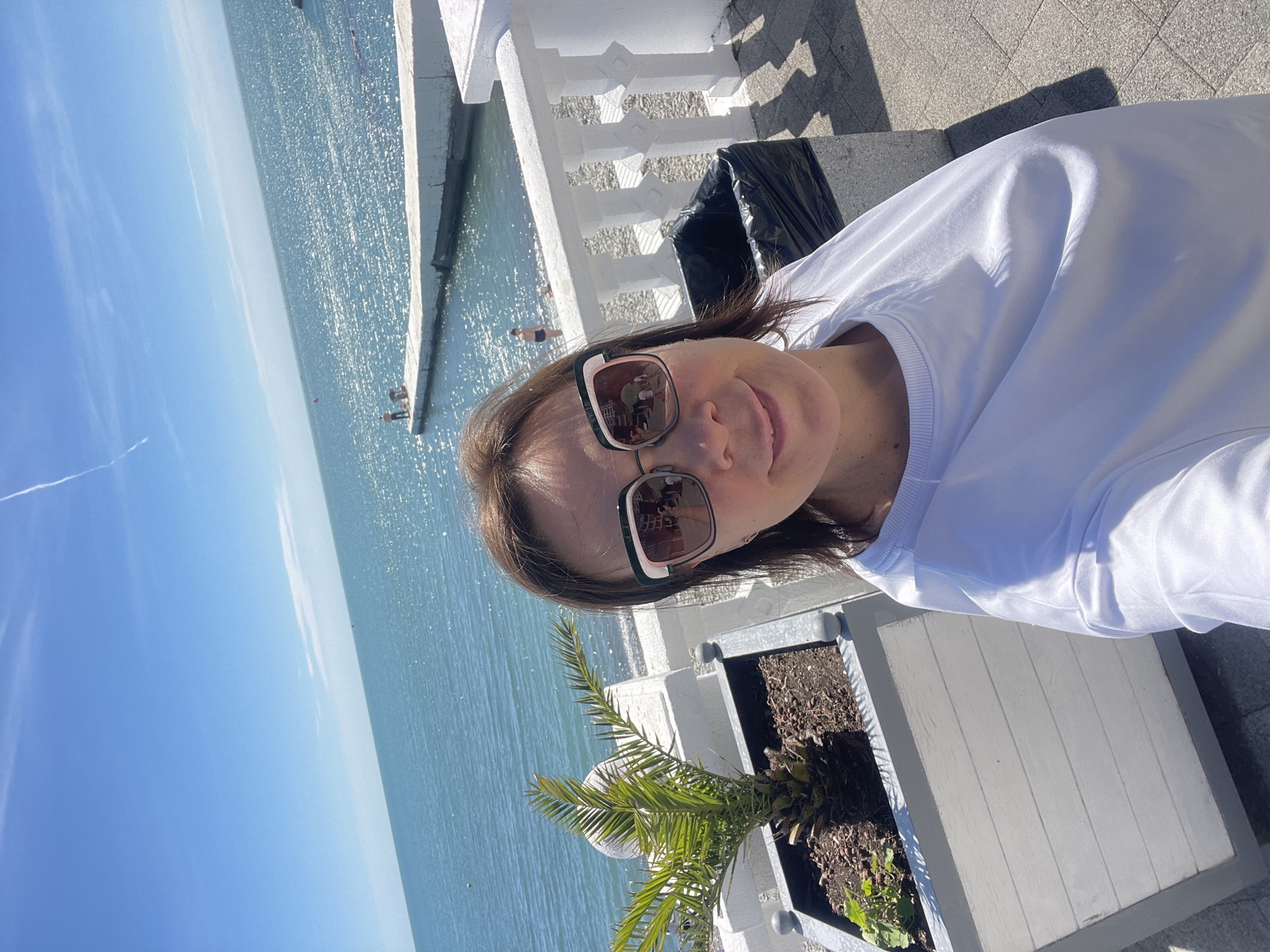 This tilted selfie captures a woman outdoors on a bright, sunny day by the sea, identified as Sochi, Russia. The woman, positioned towards the right side of the frame, has brown hair and wears light-framed sunglasses with dark lenses. She is dressed in a white long-sleeved top or sweatshirt and has a gentle smile.
Behind her, a vast expanse of calm, sparkling blue water, likely the Black Sea, meets a clear blue sky with minimal cloud cover, interrupted only by a faint contrail. Sunlight reflects vibrantly off the water's surface. In the middle distance, a concrete pier or structure extends into the sea, with a few individuals visible in the water and on the pier. A red buoy can also be seen in the water.
The foreground features a white, decorative balustrade and large, rectangular planter boxes. One planter holds a small, palm-like plant with green fronds, while another contains soil and some small green growth. A dark trash bag in a bin is partially visible next to the balustrade. The ground beneath appears to be a paved surface.
The image suggests a warm, clear day, likely midday or early afternoon, given the strong sun and clear visibility. The setting is characteristic of a seaside promenade or resort. No visible text is present.