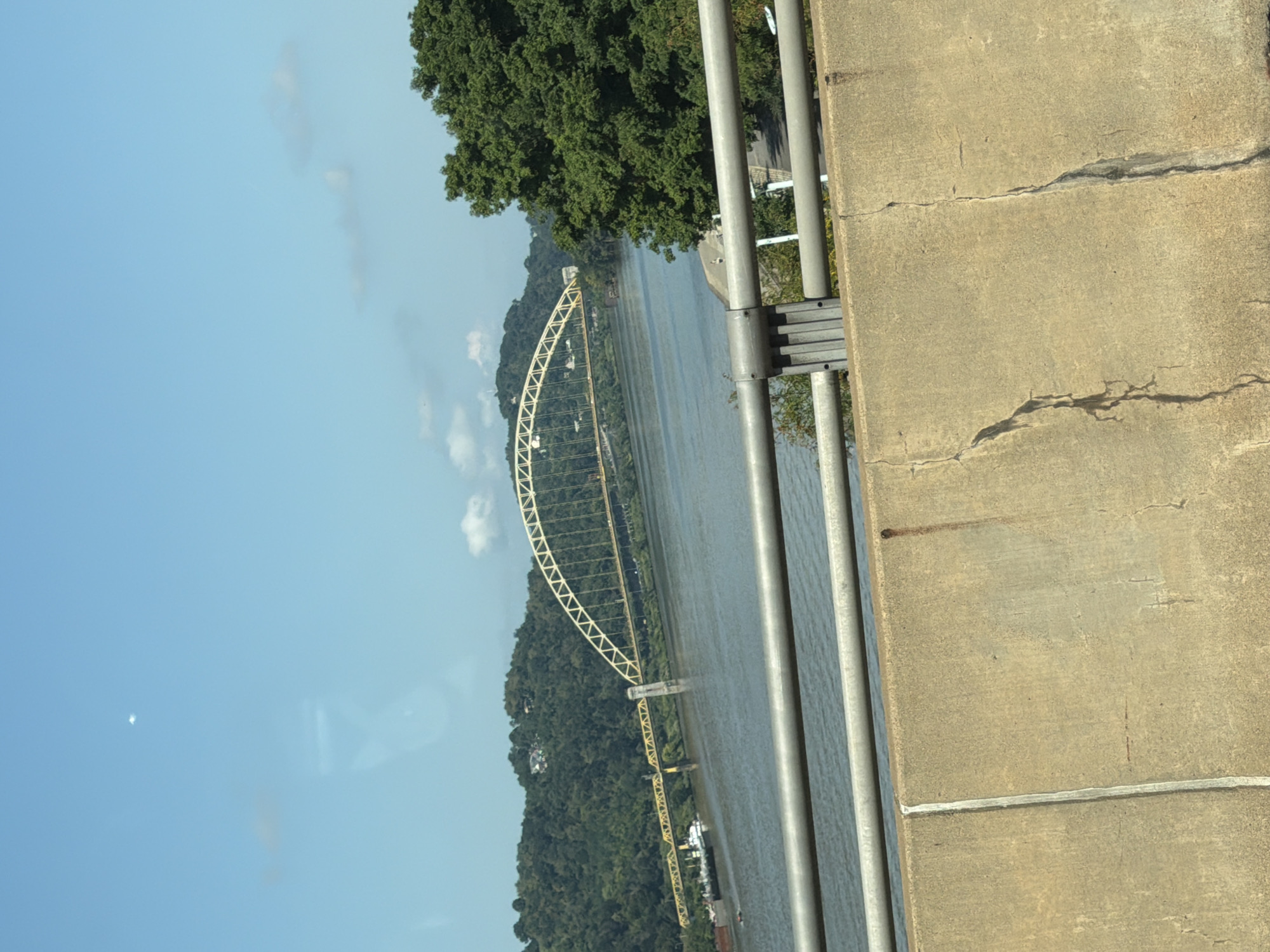 The image depicts a wide river flowing through a city, likely Pittsburgh, USA, under a clear blue sky with scattered clouds. In the background, a prominent yellow arched bridge spans the river. The riverbanks are lined with lush green trees and hills, with some buildings visible on the slopes. A few boats are present on the water. In the foreground, a concrete barrier with metal railings runs along the edge of what appears to be a roadway, suggesting the photo was taken from a vehicle or elevated vantage point. The concrete barrier exhibits visible cracks and stains.