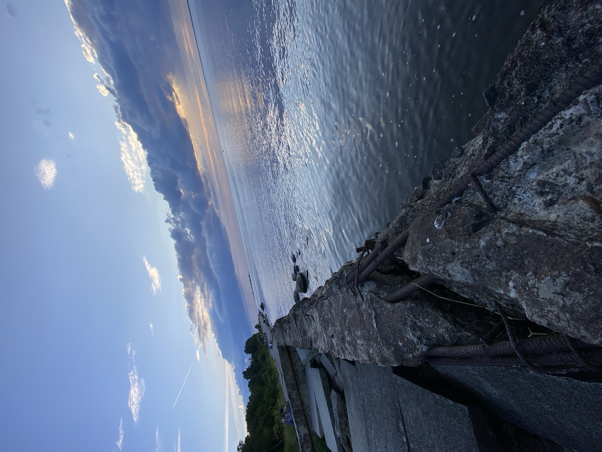 The image depicts a concrete embankment along the Gulf of Finland in Sestroretsk, Russia. The embankment is in a state of disrepair, with large concrete slabs and exposed rusted rebar visible. The sea stretches out towards the horizon, reflecting the soft light of the setting sun. Dark, heavy clouds are present in the sky, with patches of lighter clouds allowing the golden hues of the sunset to peek through. A contrail from an airplane is visible in the upper left quadrant of the sky. On the grassy area behind the embankment, a few individuals are seated or standing, observing the seascape. The overall scene is one of a somewhat dilapidated but picturesque coastal area during twilight.