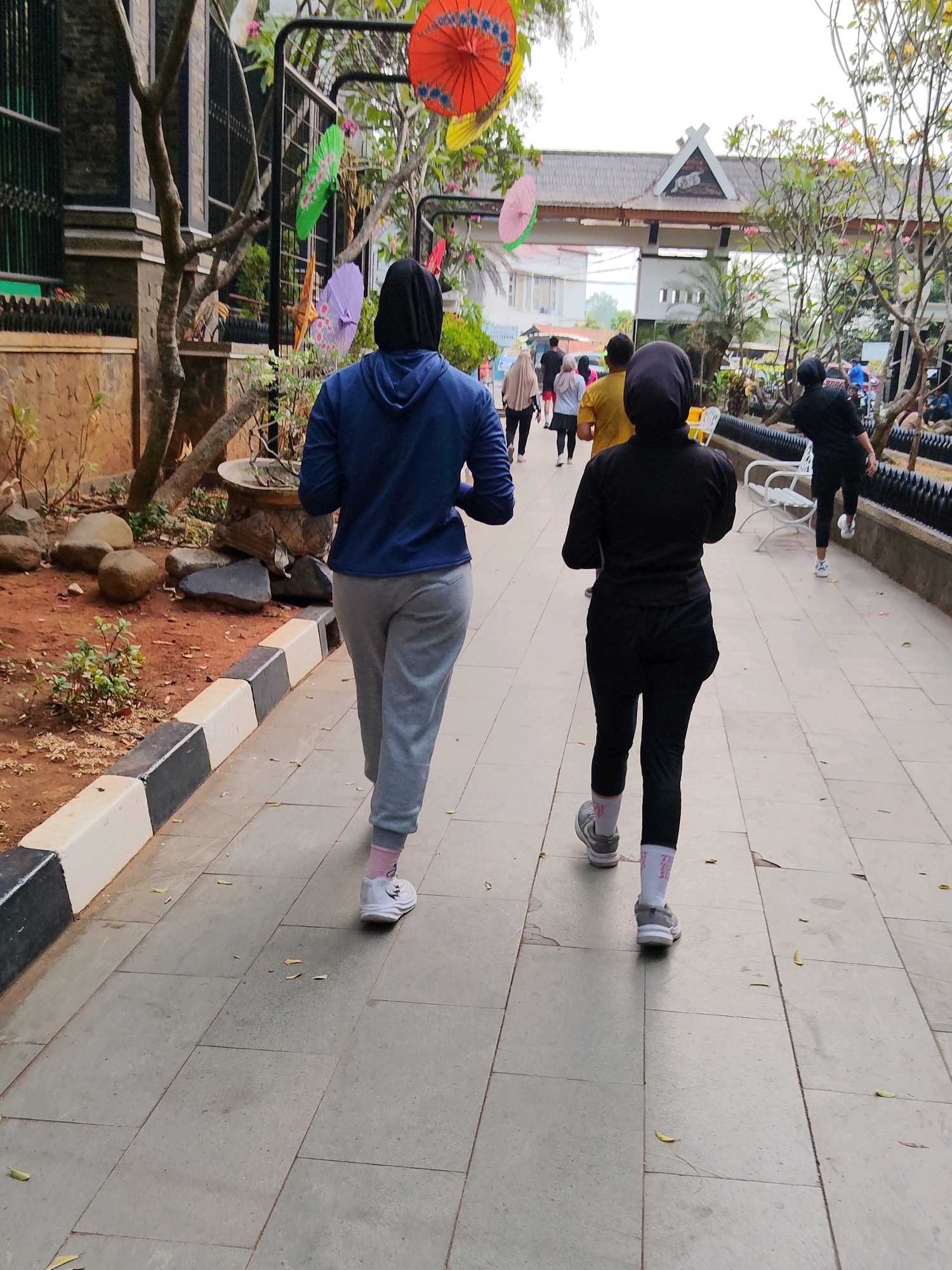 The image captures a street scene in Purwakarta, Indonesia, focusing on two individuals walking away from the camera on a paved pathway. The pathway is bordered by a decorative curb and vegetation on the left, and a railing and benches on the right. Overhead, colorful paper umbrellas are suspended from trees, adding a festive touch to the environment.
The two primary subjects are women, both wearing athletic attire and hijabs. The woman in the foreground on the right wears a black, long-sleeved top and black leggings, with grey athletic shoes and white socks. The woman in the middle ground on the left is dressed in a blue hooded jacket and grey sweatpants, also wearing athletic shoes and pink-tinged socks.
Several other individuals are visible in the background, also appearing to be engaged in recreational activity. A woman in a beige hijab and light-colored pants is walking ahead of the primary subjects, followed by a woman in a light purple hijab and white top. Further back, a group of people are seen walking towards the ornate gateway that marks the end of the visible pathway. The gateway appears to be a traditional architectural structure with a prominent roof.
The environment suggests a public park or recreational area. The ground is paved with large, square tiles. The vegetation includes trees with sparse foliage and scattered leaves on the ground. The overall atmosphere is one of casual outdoor activity, possibly a morning or evening stroll or jog, given the attire of the individuals. The lighting suggests it is daytime, but the sky is overcast.