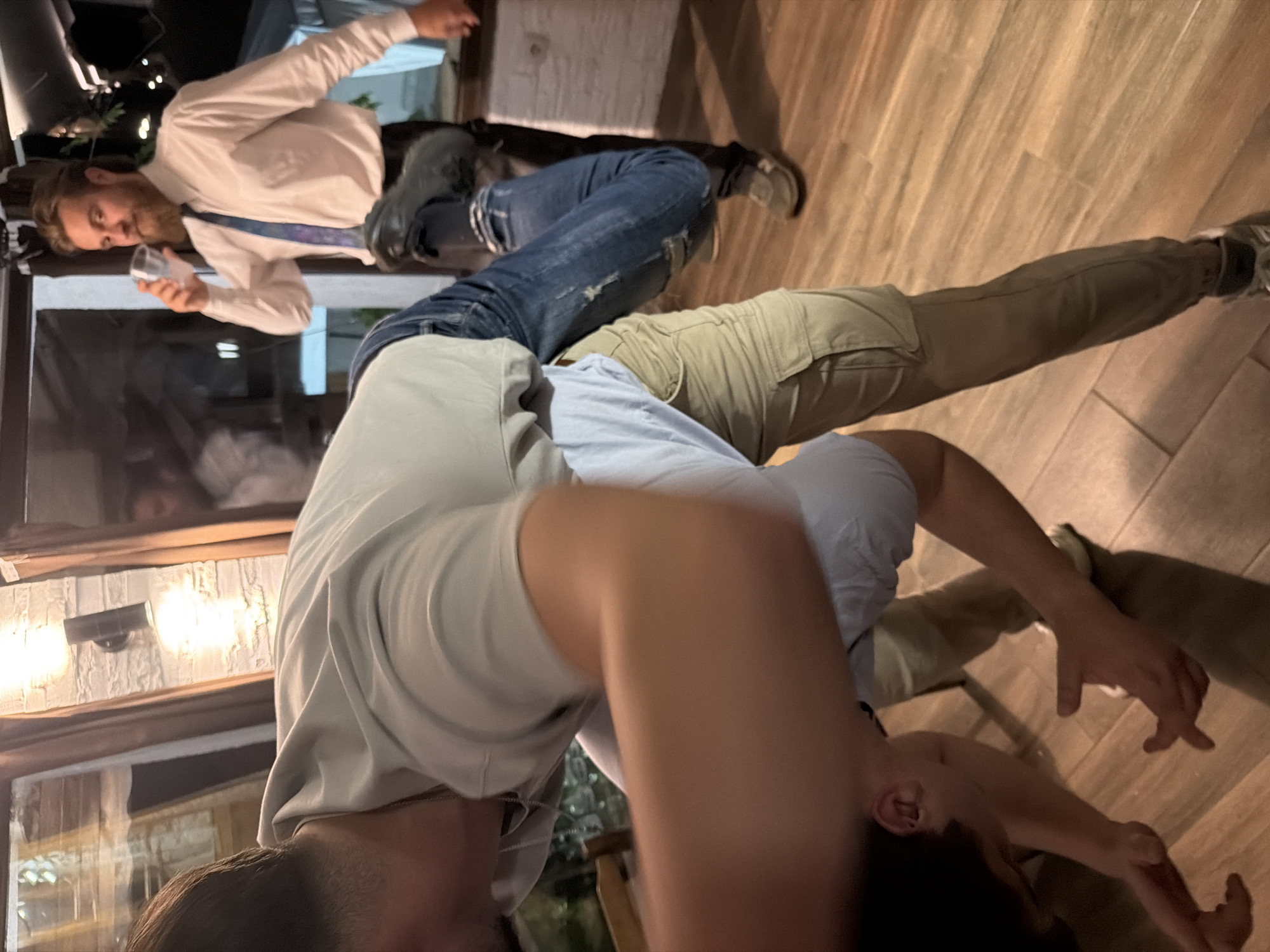 Two men are indoors. One man is wearing a white dress shirt, a blue tie, and jeans, and is holding a clear plastic cup. The other man is wearing a grey shirt and tan pants. The location appears to be a restaurant, with dark furniture, and lights on the walls. The exact location within Beli Osam, Bulgaria cannot be determined from the image.