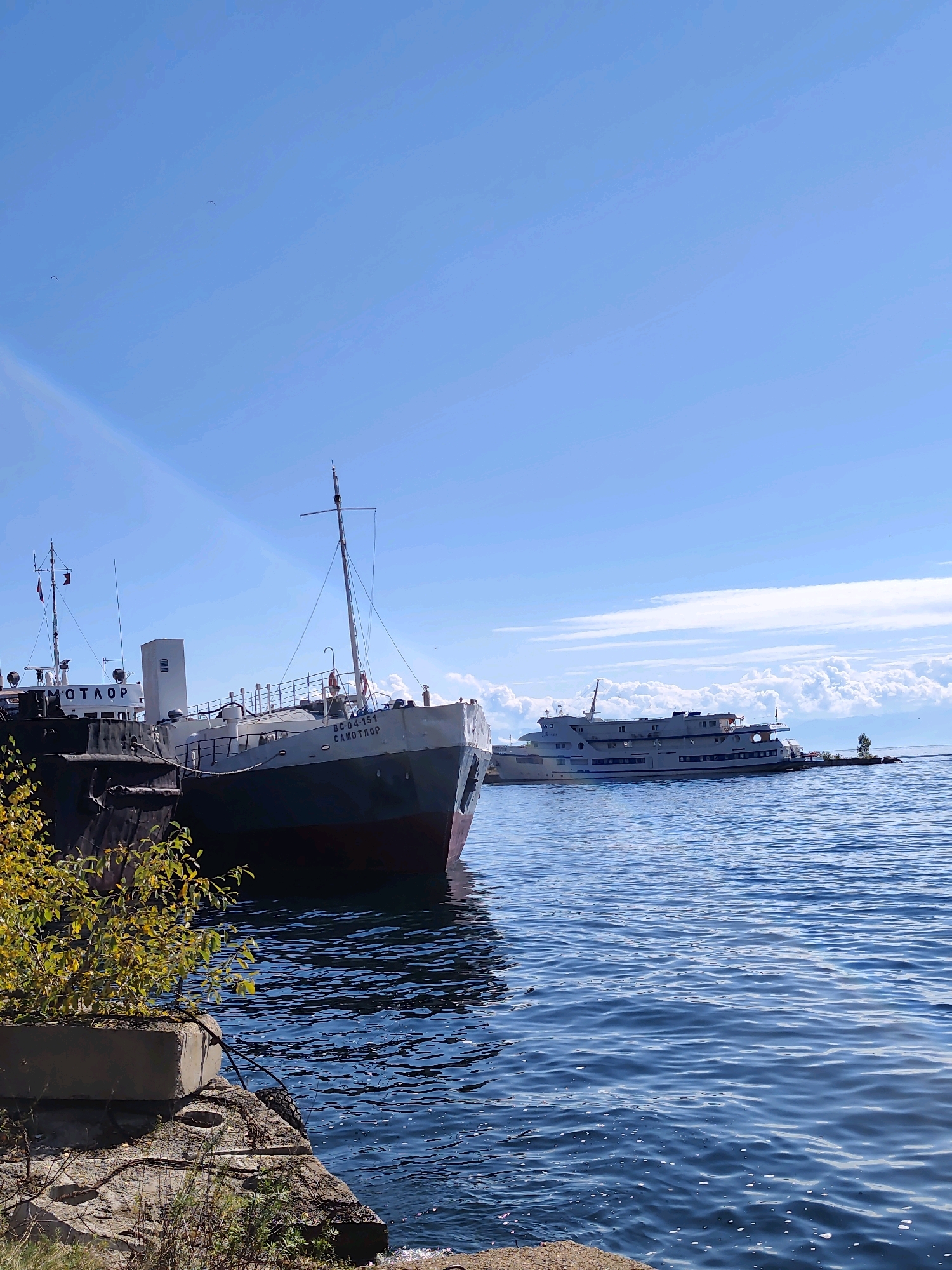 In the city of Baikal, Russia, two large ships are docked on a body of water under a clear blue sky with scattered white clouds. The ship in the foreground, partially obscured by foliage, has the name "САМОТЛОР" painted on its side. Another ship, larger and longer, is docked further in the background. The water is a deep blue and appears calm with gentle ripples. The scene depicts a peaceful harbor environment.