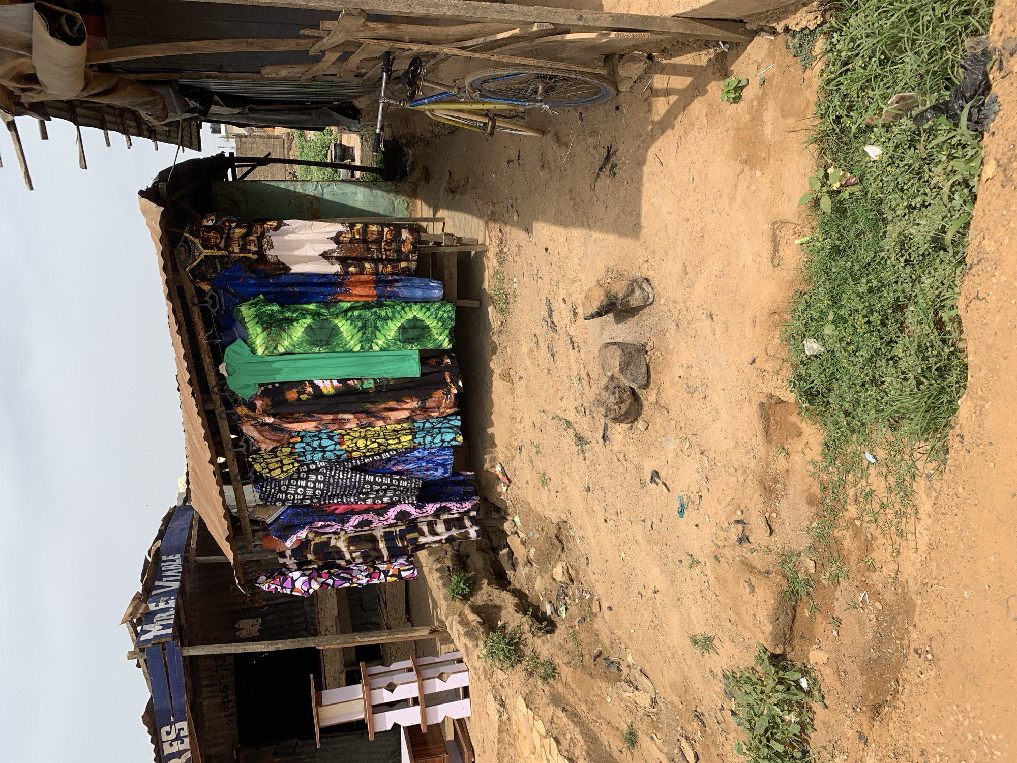 An outdoor market stall displays stacks of colorful textiles in Rafin Par, Nigeria. The stall, constructed from corrugated metal and wood, has a blue sign with white lettering, partially obscured, reading "Mr. E. Viable". A bicycle rests against the structure. The ground is dirt with patches of grass and scattered debris. There are small, dark objects on the ground near the stall.