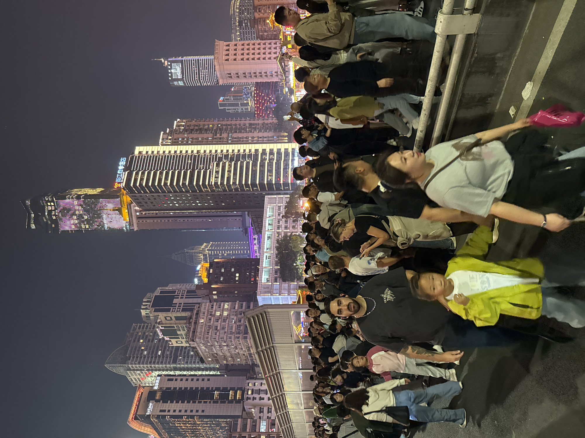 A large crowd of people, mostly young adults and children, is gathered on an elevated walkway or bridge, likely in the Yuzhong District of China, given the prominent presence of modern high-rise buildings with illuminated signs and architectural details characteristic of urban China. It's nighttime, and the skyline is brightly lit, providing a backdrop for the scene. People are looking toward the buildings or perhaps an event occurring nearby. Some individuals are taking pictures with their phones. The bridge has a metal railing.