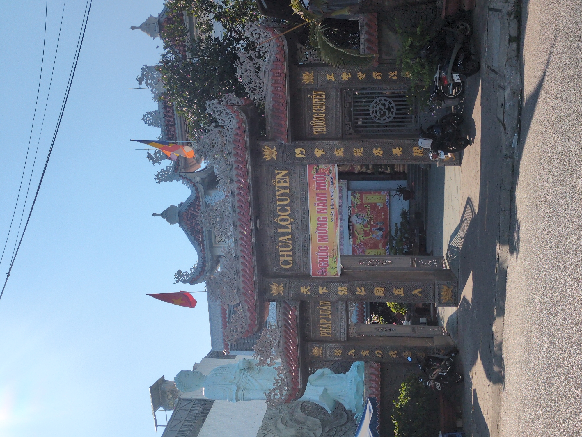 The image shows the entrance gate of the Chùa Lộc Uyển temple, located in South Quy Nhon Ward, Vietnam. The gate is ornate, featuring traditional Vietnamese architectural elements, including tiered roofs, intricate carvings, and gilded lettering of the temple's name "Chùa Lộc Uyển." A sign with the text "CHÚC MỪNG NĂM MỚI XUÂN BÌNH NGO-2026" is displayed on the gate. A statue is visible to the left of the gate. Flags fly above the entrance. Two motorbikes are parked near the entrance gate. A paved road and sidewalk are visible in the foreground. Overhead power lines are visible in the sky.
