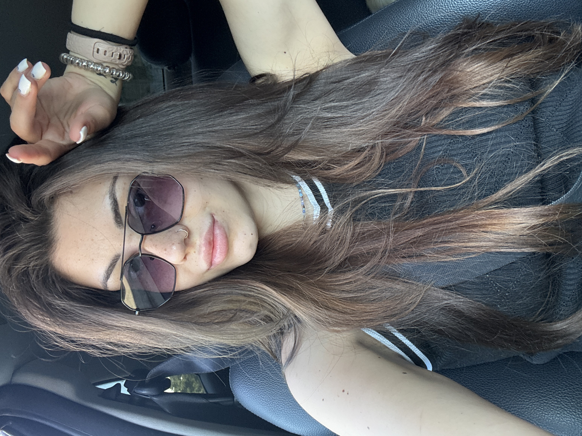 In the city of Sofia, Bulgaria, a woman with long, dark brown hair and light skin is shown in a selfie taken from inside a car. She is wearing sunglasses with dark lenses and a black top with white stripes on the shoulders. Her right hand is raised to her head, and she has white nail polish. She is also wearing bracelets on her wrist and a nose ring. The interior of the car is visible, including the black leather seat and the seatbelt. The lighting suggests it is daytime, possibly late afternoon, with sunlight filtering into the vehicle. The background outside the car appears to be blurred foliage, indicating an outdoor setting, possibly a street or a park.