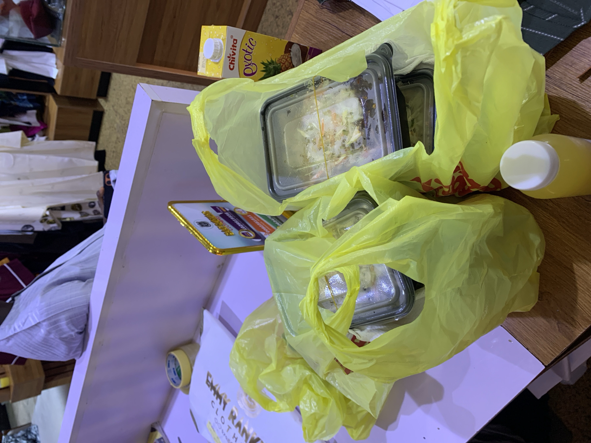 The image shows a close-up of several yellow plastic bags on a wooden table, containing food in plastic containers. To the right of the bags is a carton of Chivita Exotic juice. In the background, a person is visible wearing a light-colored robe, and in the further background, there are clothing racks with garments. The scene appears to be indoors, possibly a shop or a home, with fluorescent lighting. The presence of the food and juice suggests a recent meal or a prepared order. The overall impression is one of a casual, everyday setting in Gura Top, Nigeria.