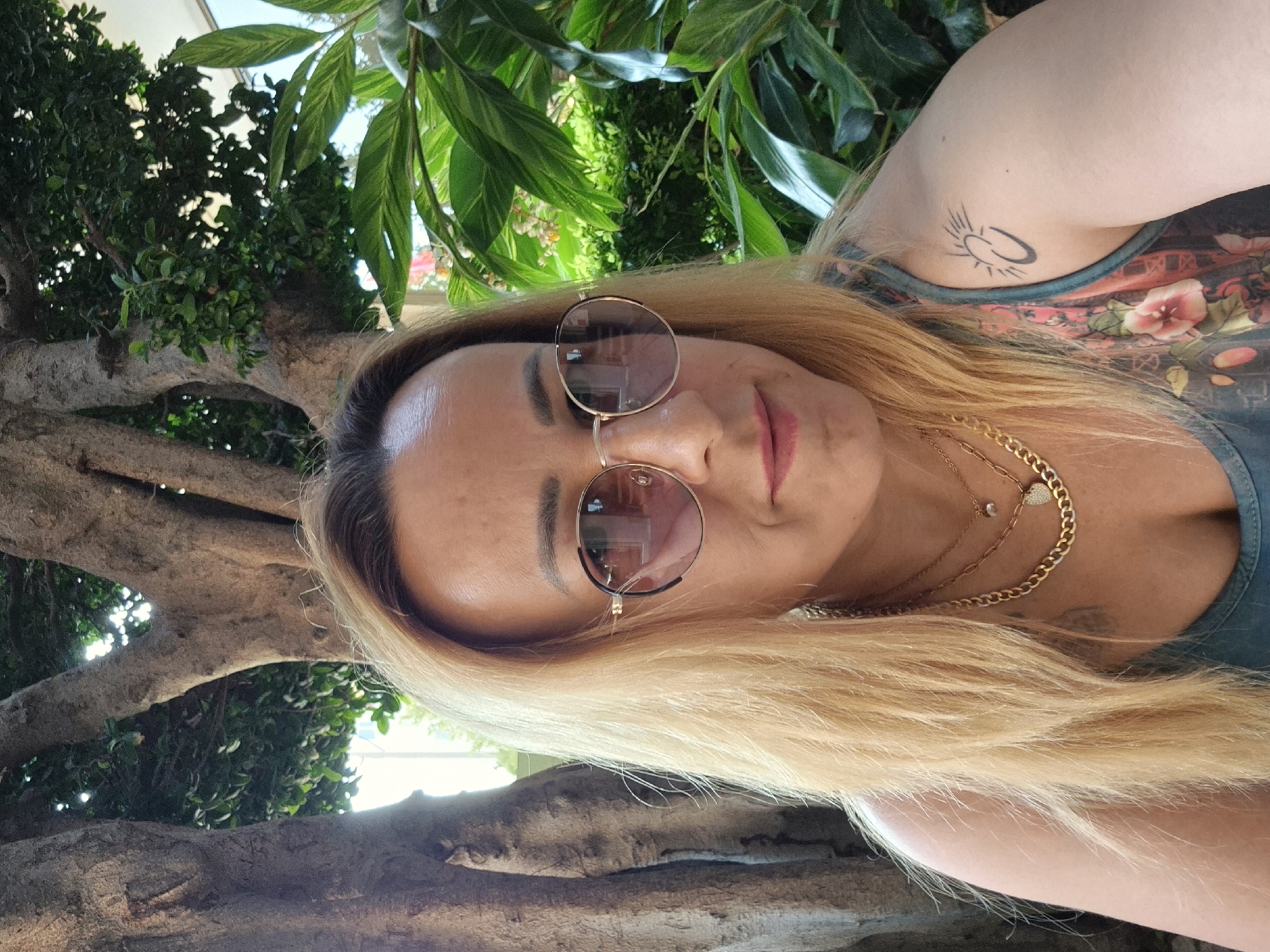 An outdoor, waist-up, medium shot captures a fair-skinned blonde woman with light brown eyebrows, a light dusting of freckles, and pink lipstick, looking at the camera in the city of Unknown, Unknown. She is wearing round, gold-rimmed sunglasses with pink lenses, and a dark teal tank top with a floral print of pink and red flowers. She has on two gold necklaces; the top one is thin with a small, clear pendant, and the bottom one is a thicker chain with a heart-shaped pendant. Her hair is light blonde and falls to her shoulders. On her left shoulder, there is a black tattoo of a sun with lines radiating from it. Behind her, to the left, is a large tree trunk with rough, gray bark. To the right of the trunk is lush, green foliage with large leaves and some smaller yellow flowers. The lighting suggests it is daytime, with sunlight filtering through the leaves.
