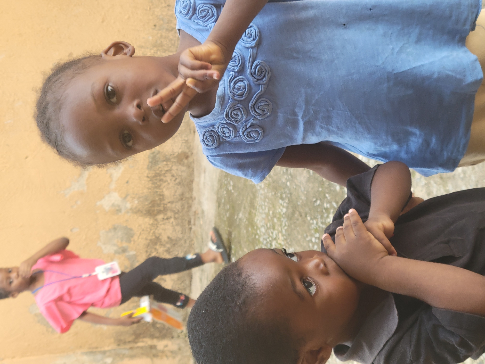The image depicts three children in an outdoor setting. One child, wearing a blue shirt with floral appliqués, is holding up a "peace" sign. Another child, wearing a black shirt, is holding their hands near their face. In the background, a third child, wearing a pink shirt and black pants, is partially visible, holding a yellow and orange object. The background suggests an urban environment, with what appears to be a building wall and paving, potentially in Suleja, Nigeria. The photo has been rotated.