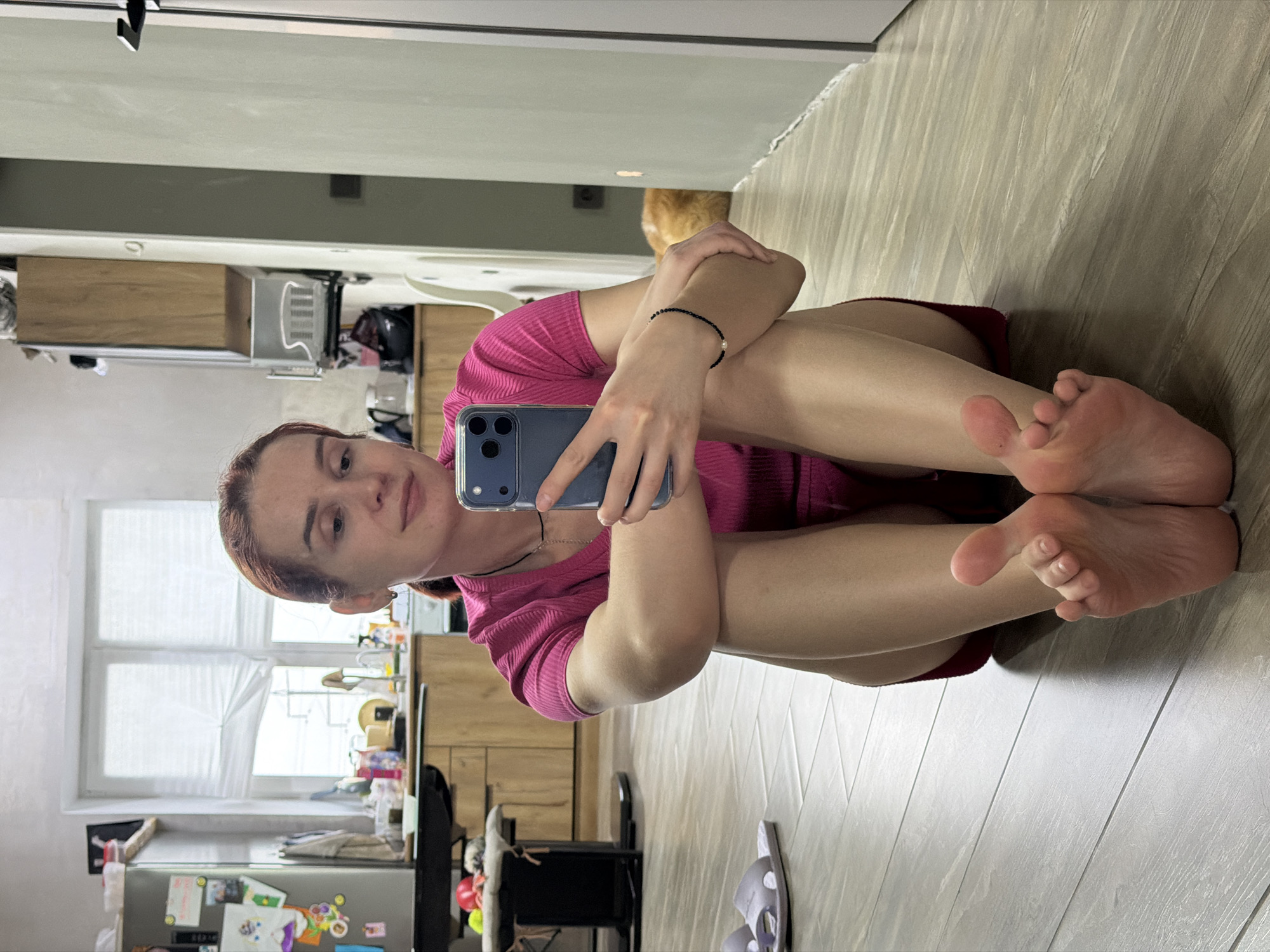 A woman is sitting on a light-colored wooden floor, indoors. She is fair-skinned with reddish-brown hair, wearing a pink shirt, and holding a smartphone up to take a selfie. Her legs are bent and pulled close to her torso. A glimpse of her bare feet is visible. The background reveals a kitchen space with a window, refrigerator, and cabinets. There is no indication this is in the city of Chaikovsky, Russia.