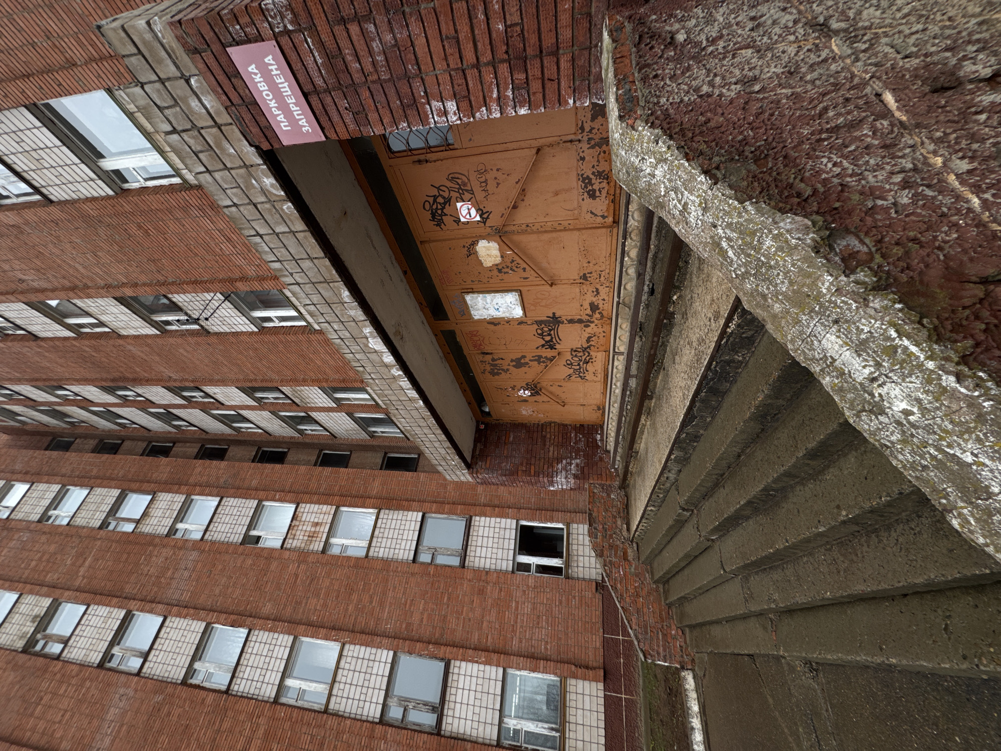 The image depicts an outdoor urban scene in Votkinsk, Russia. A multi-story brick building with multiple windows arranged in horizontal rows stands to the left. A concrete staircase runs vertically along the right side of the frame. An overhead metal structure with graffiti on it extends out above the stairs. A sign with text in Cyrillic script is visible above the metal structure on the brick building. The photograph is taken from a low angle, looking upward, giving the impression of height and verticality. The overall scene appears to be of an aged, perhaps abandoned, building.