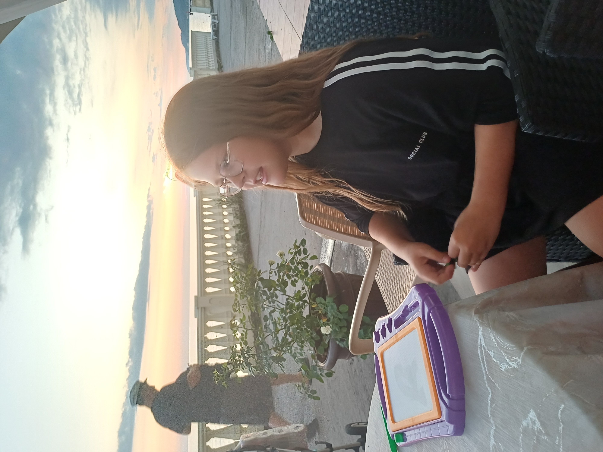 A medium shot captures a young girl with long brown hair and glasses sitting at a table outdoors, facing to the right. She wears a black t-shirt with white stripes down the sleeves and "SOCIAL CLUB" printed on the chest. In front of her is a purple and orange drawing toy with a blank screen. To her left, a person wearing a dark shirt and a light-colored hat walks away from the camera, towards a low white railing. Behind the railing is a body of water, likely the Black Sea, with a beach and mountains visible in the distance. The sky is a soft gradient of orange, yellow, and blue, suggesting it is either sunrise or sunset. The environment appears to be a promenade or a cafe terrace in Sochi, Russia, known for its coastal scenery and resorts. The lighting is warm and diffused.