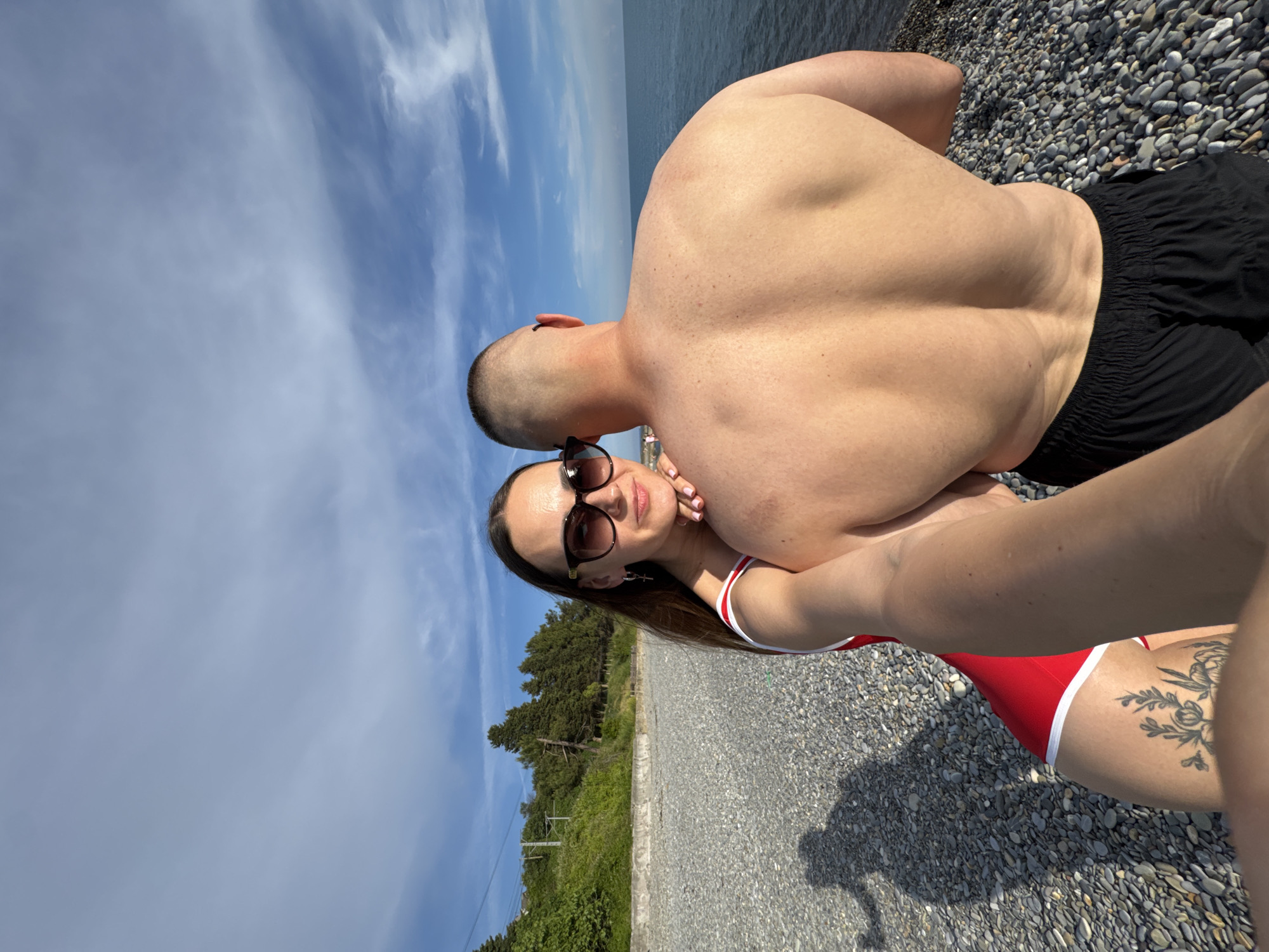 A couple is taking a selfie on a pebble beach in Sochi, Russia. The woman is in the foreground, wearing a red and white swimsuit and sunglasses, with her arm around the man. The man's back is to the camera, showing his muscular physique. The ocean is visible in the background, along with a line of trees and a blue sky with clouds. A shadow of the couple is cast on the beach.