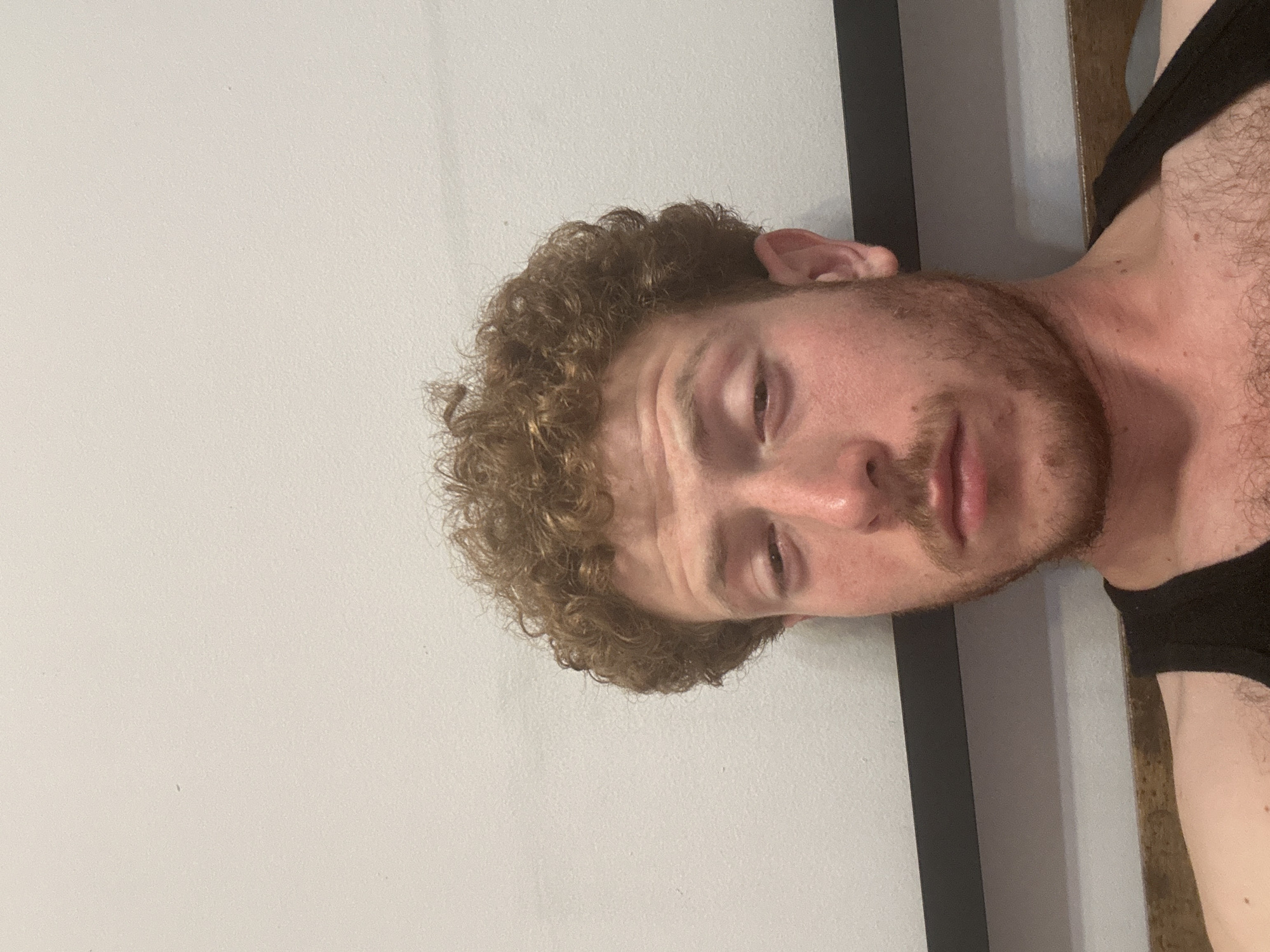A man with fair skin, curly light brown hair, and a beard is the central subject. He is wearing a black tank top. He appears to be indoors, positioned against a plain white wall and what appears to be a wooden door frame with a black accent. The environment is simple and undefined. There is no evidence in the photograph to indicate that the image was taken in New York City or any other specific location. The image provides no information about actions, interactions, or specific objects within the environment.