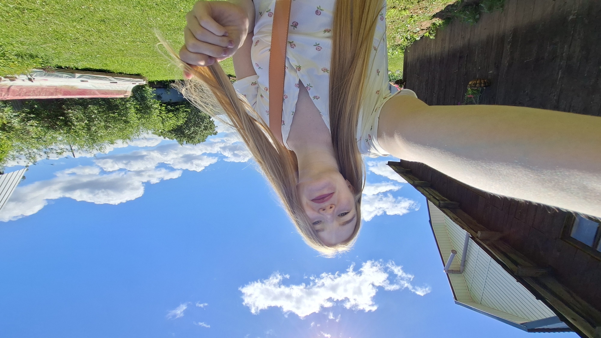 A young woman with long blonde hair and light-colored clothing takes a selfie outdoors in the village of Bolshoy Bukor, Russia. She is standing in a grassy yard with a wooden building to her left and lush green trees and bushes in the background. A bright blue sky with white clouds and the sun shining through are visible. She is holding a portion of her hair with her right hand.