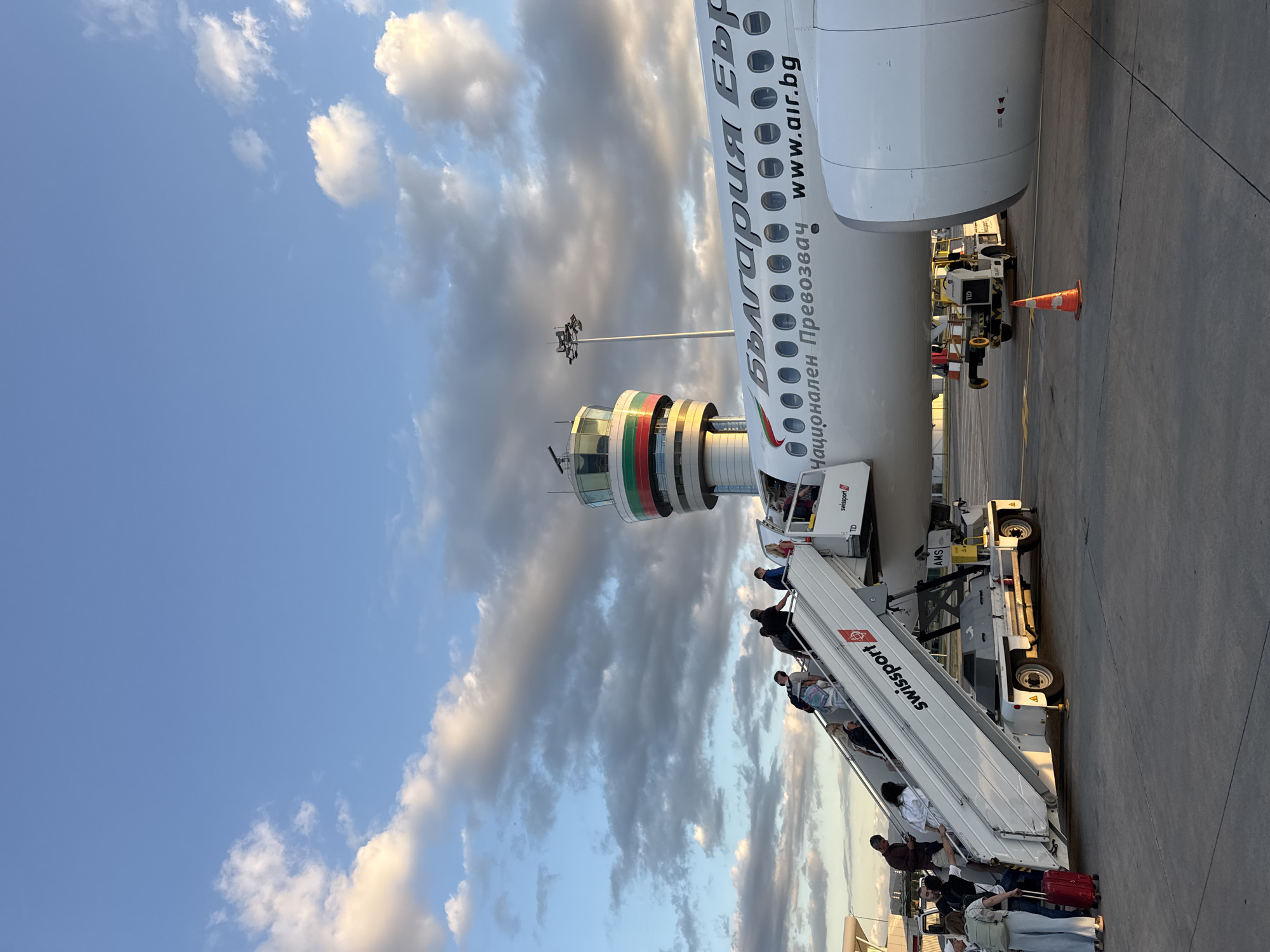 The image depicts an outdoor scene at an airport, likely Sofia Airport in Bulgaria, as indicated by the text on the airplane. Passengers are boarding a large white airplane via a portable set of stairs. The stairs are white with red accents and have the "Swissport" logo visible. Several individuals are ascending the stairs, some carrying luggage. The airplane is branded with Cyrillic text that appears to read "България Ер" and "Национален Превозвач", with "www.air.bg" also visible, suggesting it is a Bulgarian airline. In the background, a distinct airport control tower with red and green bands is visible, along with a lamppost with lights. The sky is partly cloudy with a warm, late-afternoon light casting shadows. The tarmac is visible in the foreground, with some airport service vehicles and a traffic cone.