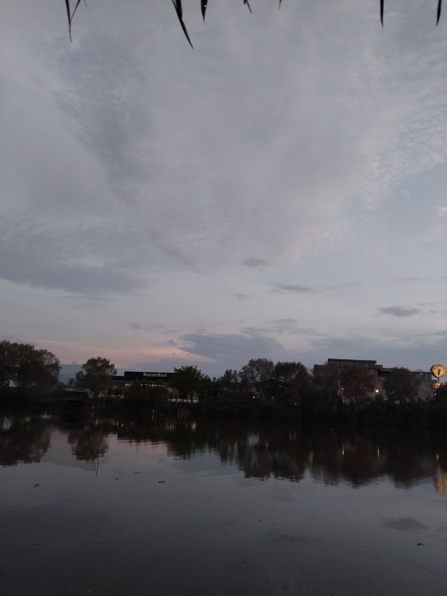 The image depicts a tranquil body of water in Lhokseumawe, Indonesia, reflecting the twilight sky and a silhouetted treeline. In the background, a building with the sign "Harun Mart" is visible, illuminated by lights. The sky is overcast with soft clouds, hinting at the end of the day. The foreground is dominated by the dark water, with subtle ripples and reflections of the surrounding scenery. The overall atmosphere is calm and serene, characteristic of a peaceful evening in the city.
